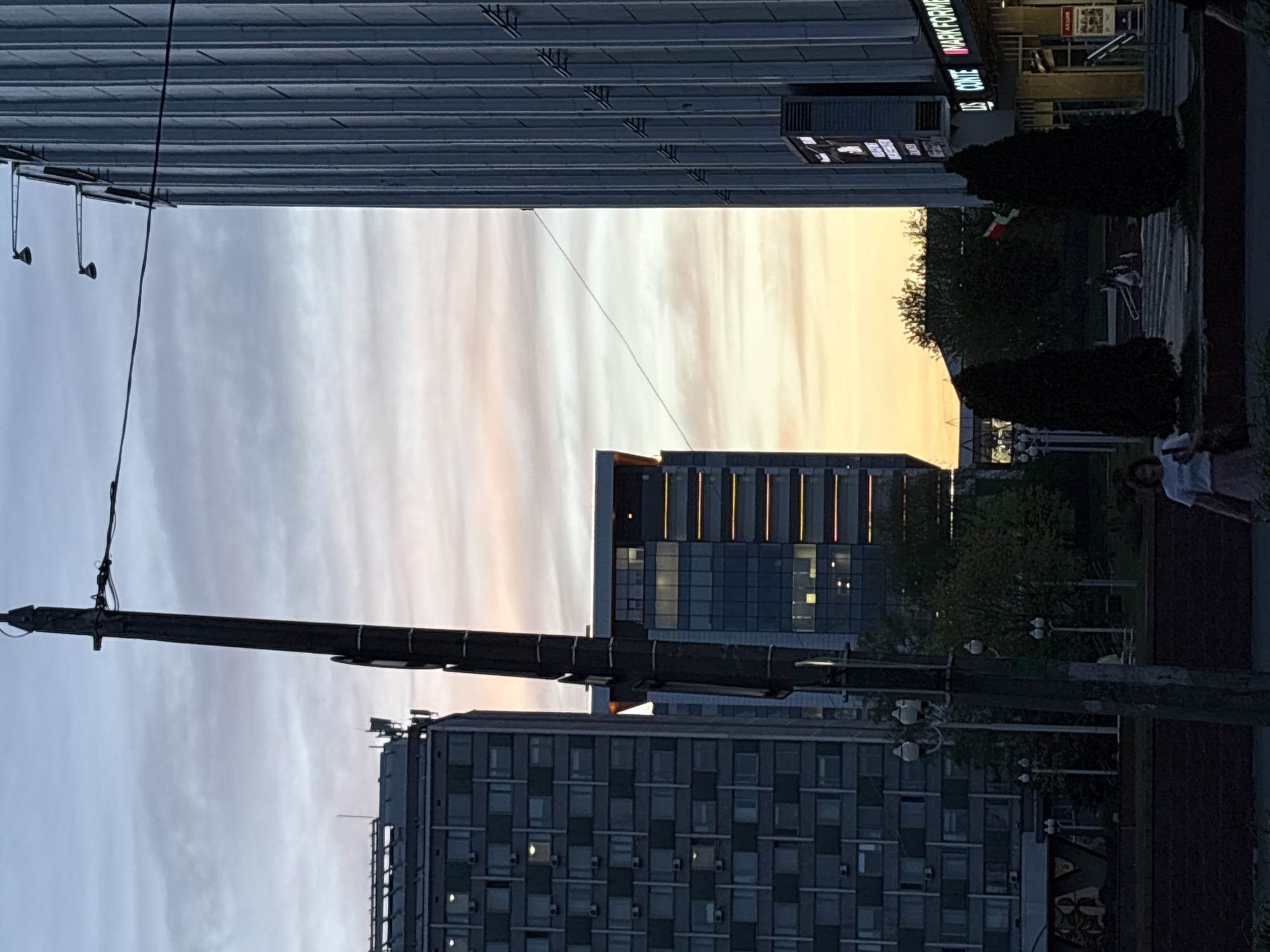 The image depicts an urban scene in Minsk, Belarus, at either sunset or sunrise, as evidenced by the orange and pink hues in the sky between buildings. A tall, gray building with numerous windows stands prominently in the foreground. In the background, another building with vertical illuminated stripes is visible. A utility pole runs across the frame, obstructing part of the view. On the right side, there are trees and a sidewalk where a person is walking. A building with the name "MARK FORMELLE" is also seen.