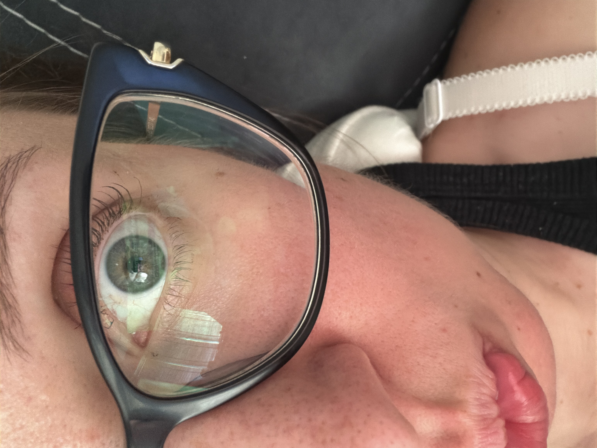 The image is a close-up, eye-level shot of a person's face, focusing on their left eye and part of their mouth, nose, and cheek. The individual is wearing dark-rimmed glasses, and their eye, which is green with brown flecks, is visible through the left lens. The reflection in the glasses shows some light sources. The skin is fair with some freckles. A white bra strap and a black garment are visible in the background, suggesting the person is wearing clothing. The image does not contain any contextual information or visual cues that would allow for the determination of its location in Bila Tserkva, Ukraine, or any urban environment.