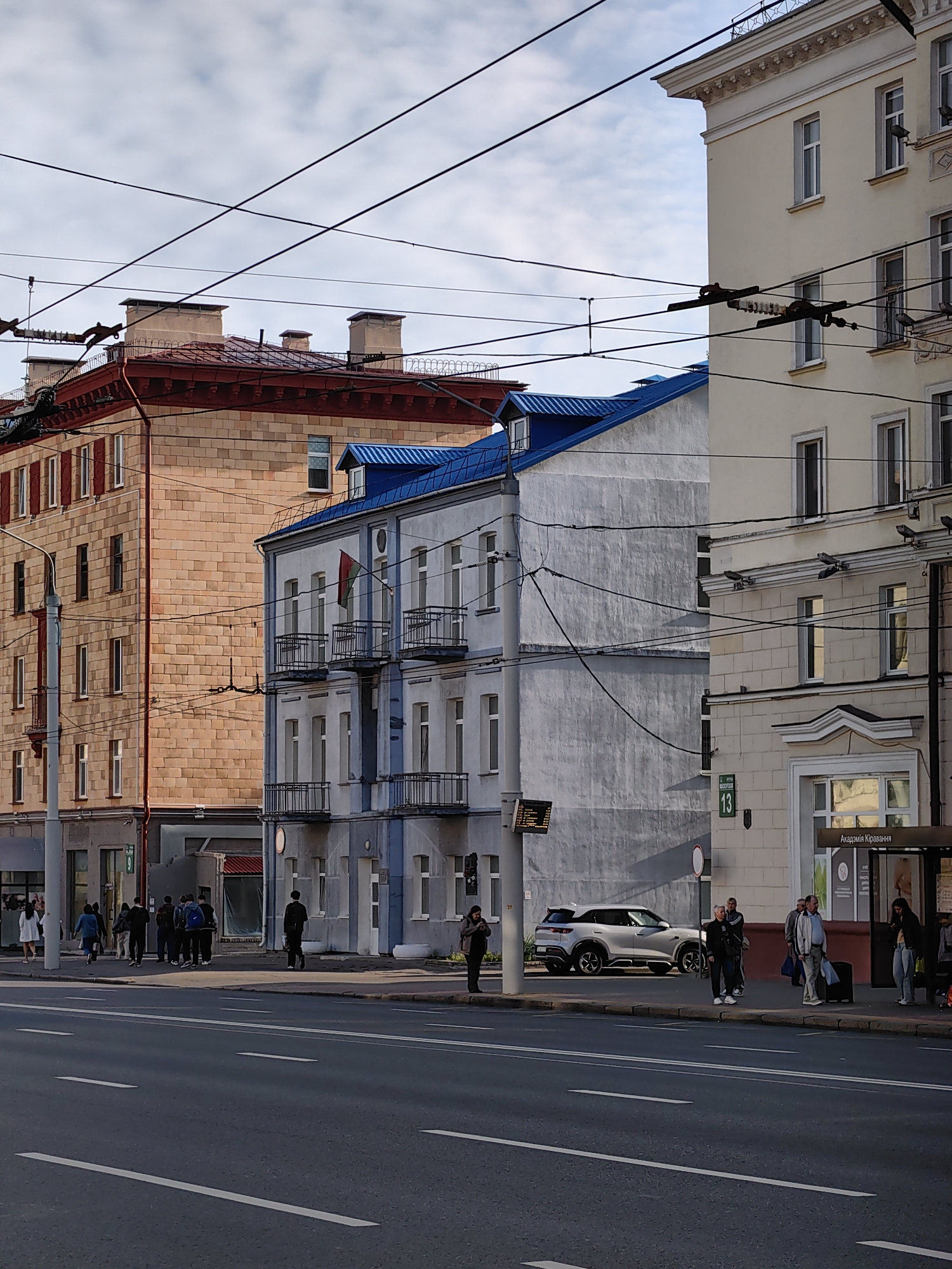 The image shows a street scene in Minsk, Belarus, likely during the daytime given the lighting. In the foreground, a multi-lane asphalt road with white lane markings is visible. On the right side of the road, a bus stop shelter with signage in Cyrillic script is present. Several people are waiting at or near the bus stop, some standing, and one person is pulling a suitcase. A light-colored modern SUV is parked on the side of the road behind the bus stop.
Behind the sidewalk, a series of buildings line the street. On the left, a multi-story building constructed with light brown bricks features multiple windows and a red roofline with decorative elements. To its right, a narrower building with a blue pitched roof is visible, with light-colored facades and balconies. Further to the right, a taller, pale yellow building with a white decorative cornice and numerous windows dominates the scene. Overhead, a complex network of overhead electrical wires, likely for trolleybuses, crisscrosses the sky above the buildings and street. The sky is partly cloudy.
On the sidewalk in front of the pale yellow building, a green street sign with the number "13" is mounted. The building facade also has signage that appears to be in Cyrillic. The general architectural style suggests an urban setting in a post-Soviet era city.