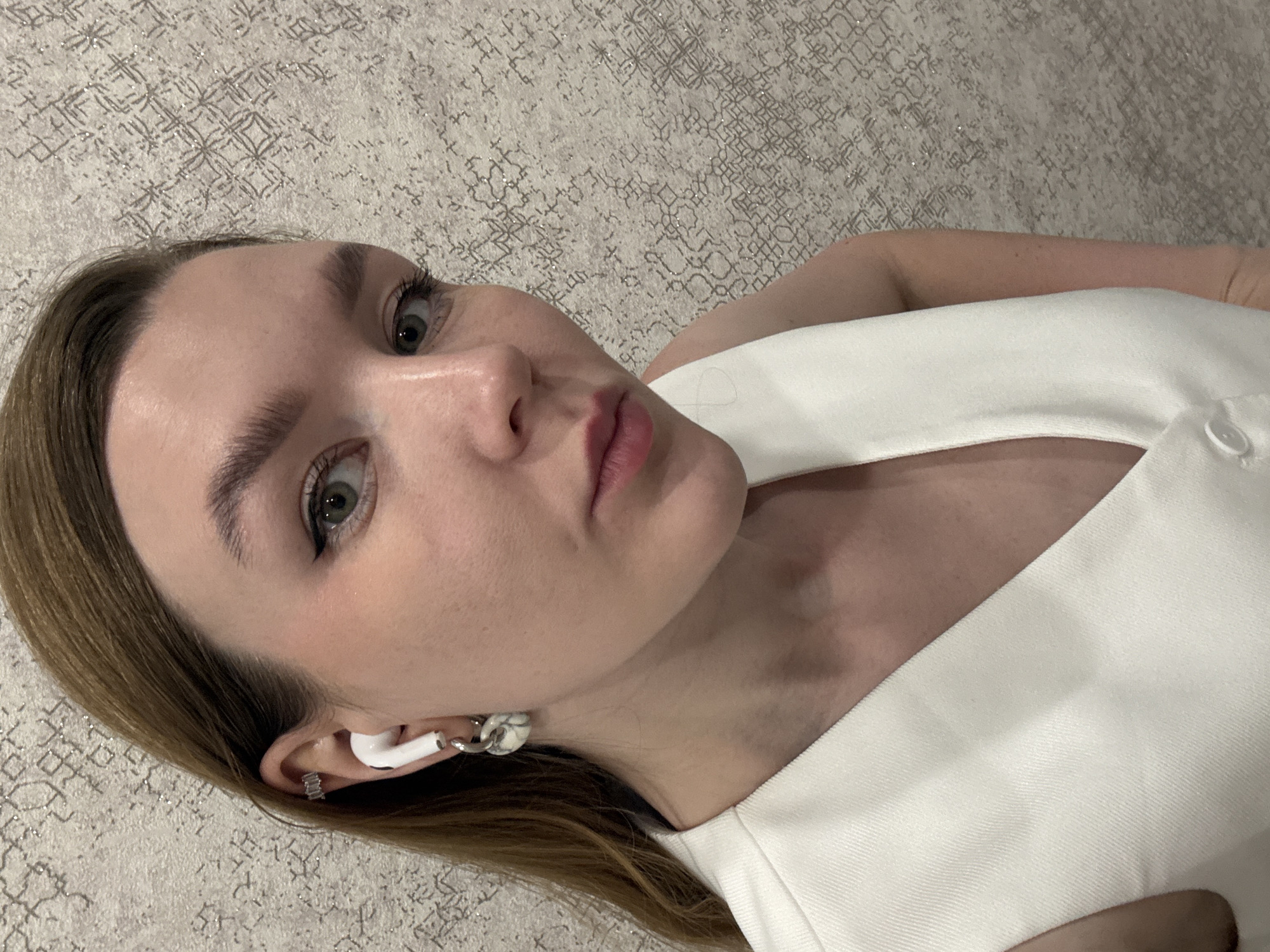 A young woman with fair skin, light brown hair, and green eyes poses for a selfie. She is wearing a white sleeveless top. Her hair is parted to the side, with some strands falling near her face. She has an earbud in one ear and wears an earring. The background appears to be a textured carpet or rug with a patterned design. The overall impression is a casual, close-up portrait, but there is no information to suggest it was taken in Izhevsk, Russia.