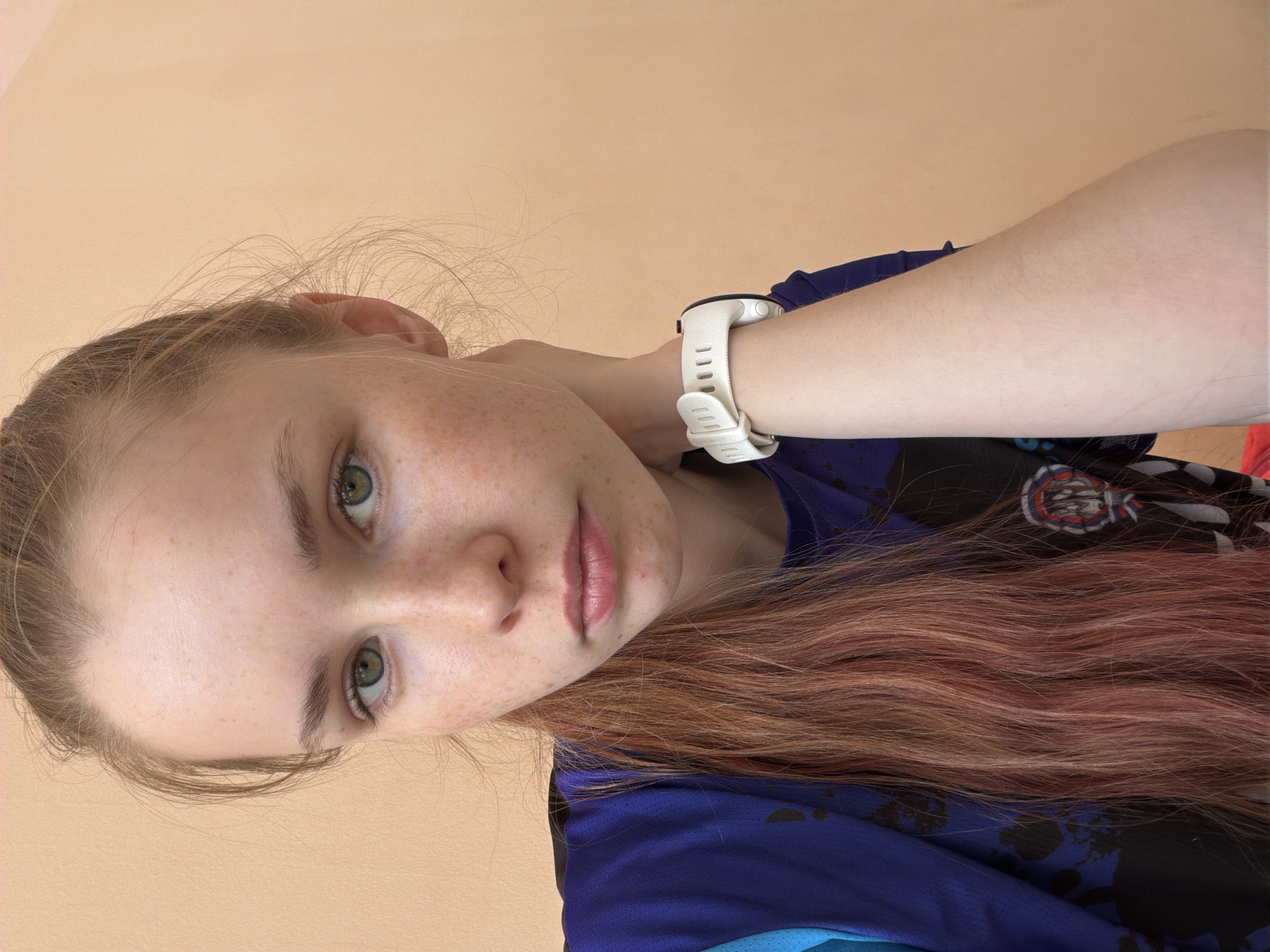 A close-up portrait shot features a young woman with fair skin and numerous freckles scattered across her nose and cheeks. She has light green or hazel eyes that gaze directly at the camera, and her expression is neutral and calm. Her hair is long, appearing light brown or blonde with noticeable reddish or pinkish undertones, and cascades over her left shoulder. She is wearing a blue short-sleeved top adorned with subtle black patterns; a partially visible white and red circular logo is present on the upper part of the shirt. A white smartwatch with a textured strap is prominently displayed on her left wrist. Her left hand is raised, with her arm bent and positioned near her neck. The background is a plain, light beige or peach-colored wall, indicating an indoor setting with soft, even lighting. The image is taken at an angle, with the subject's head tilted. No specific time of day or location cues are visible within the image itself, beyond the general indoor environment, though the image's origin is specified as Berezniki, Russia. There is no visible text on her clothing or the smartwatch.