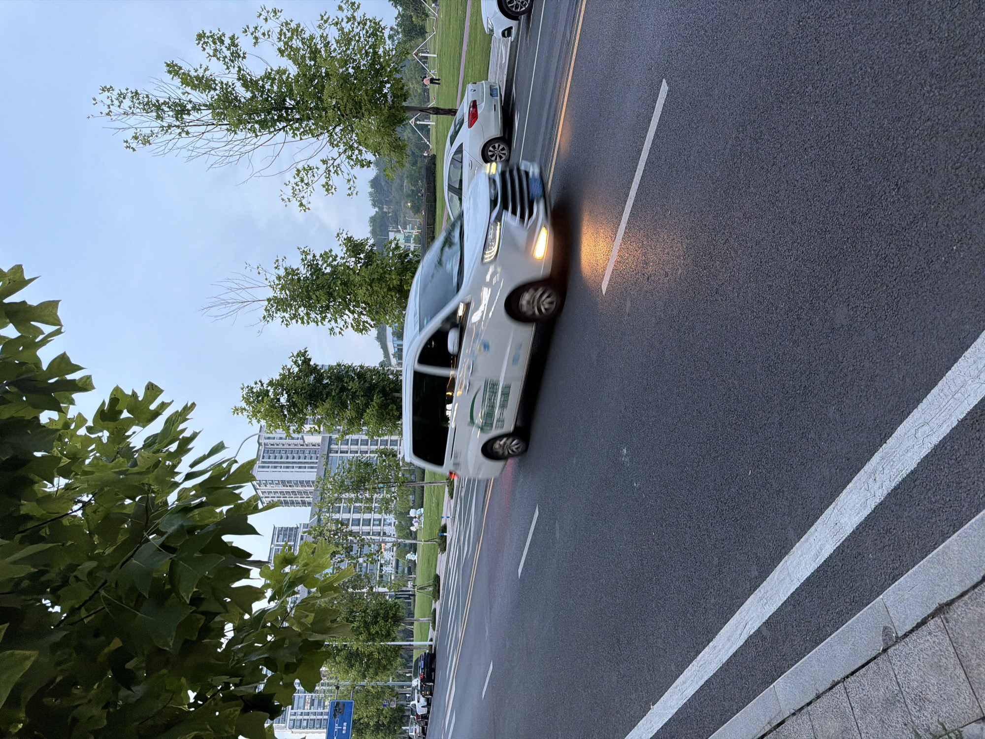 The image depicts a street scene in the city of Jiangjin, China. A white minivan, with a green logo on its side, is in motion, its headlights casting a warm glow on the asphalt road. The vehicle is captured with motion blur, indicating movement. To the right of the minivan, a white sedan is parked. The road has white lane markings. In the background, a row of tall, modern buildings stands against a pale sky. Green trees line the street and a grassy area with some playground equipment is visible on the right side of the road. The overall environment suggests a contemporary urban setting, possibly in the late afternoon or early evening, given the lighting conditions.