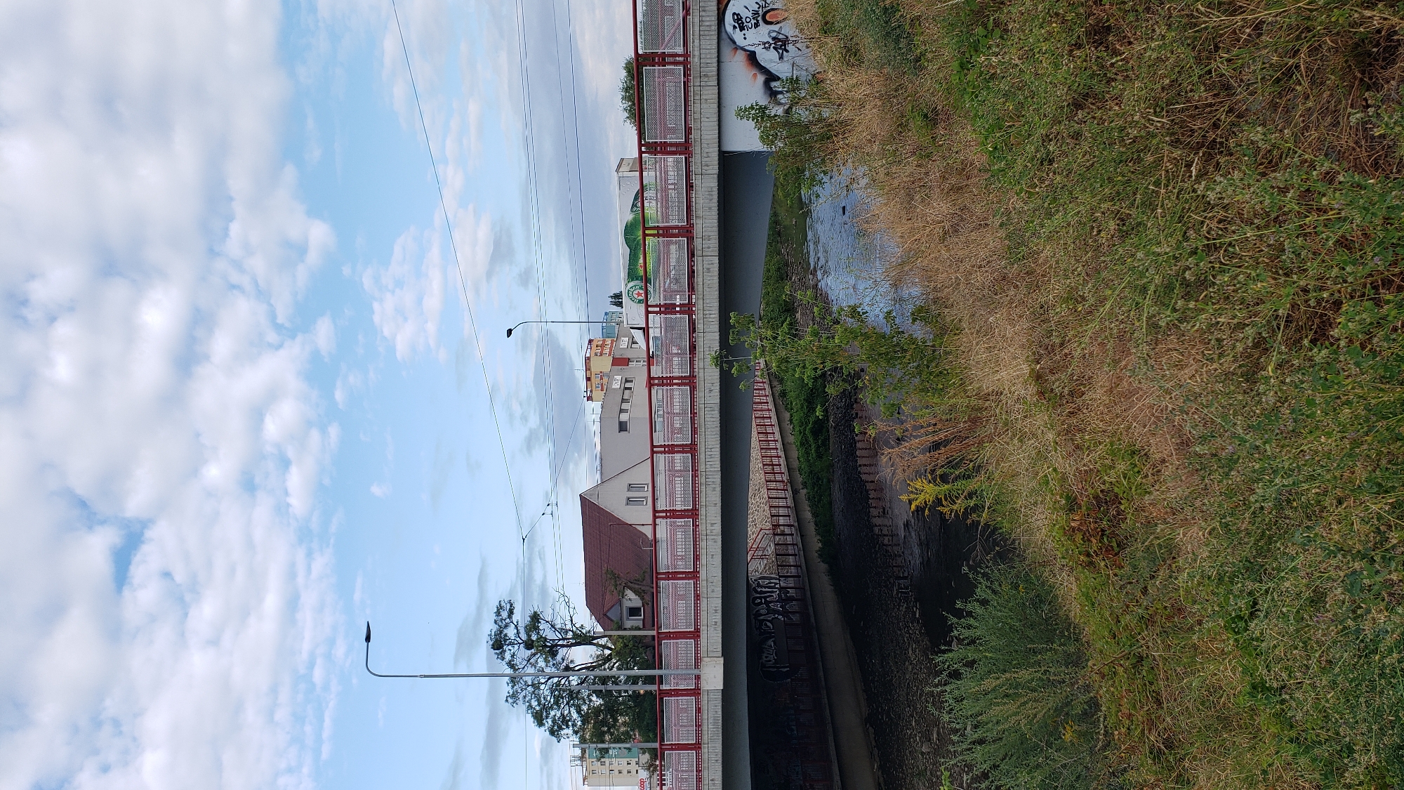 The image depicts a view of a bridge over a small body of water in Prešov, Slovakia. The bridge features a red railing with a metal mesh infill and is supported by concrete piers. Underneath the bridge, graffiti is visible on the concrete walls. On the far bank of the water, dense green and dry grass and bushes line the edge. In the background, several buildings are visible, including a red-roofed house with white walls, a taller grey building, and other structures that appear to be commercial or residential. A truck with a logo is parked near one of the buildings. Streetlights are present along the street that runs across the bridge, and power lines crisscross the sky. The sky is partly cloudy with a light blue hue. The overall scene is a typical urban landscape featuring infrastructure and nature.