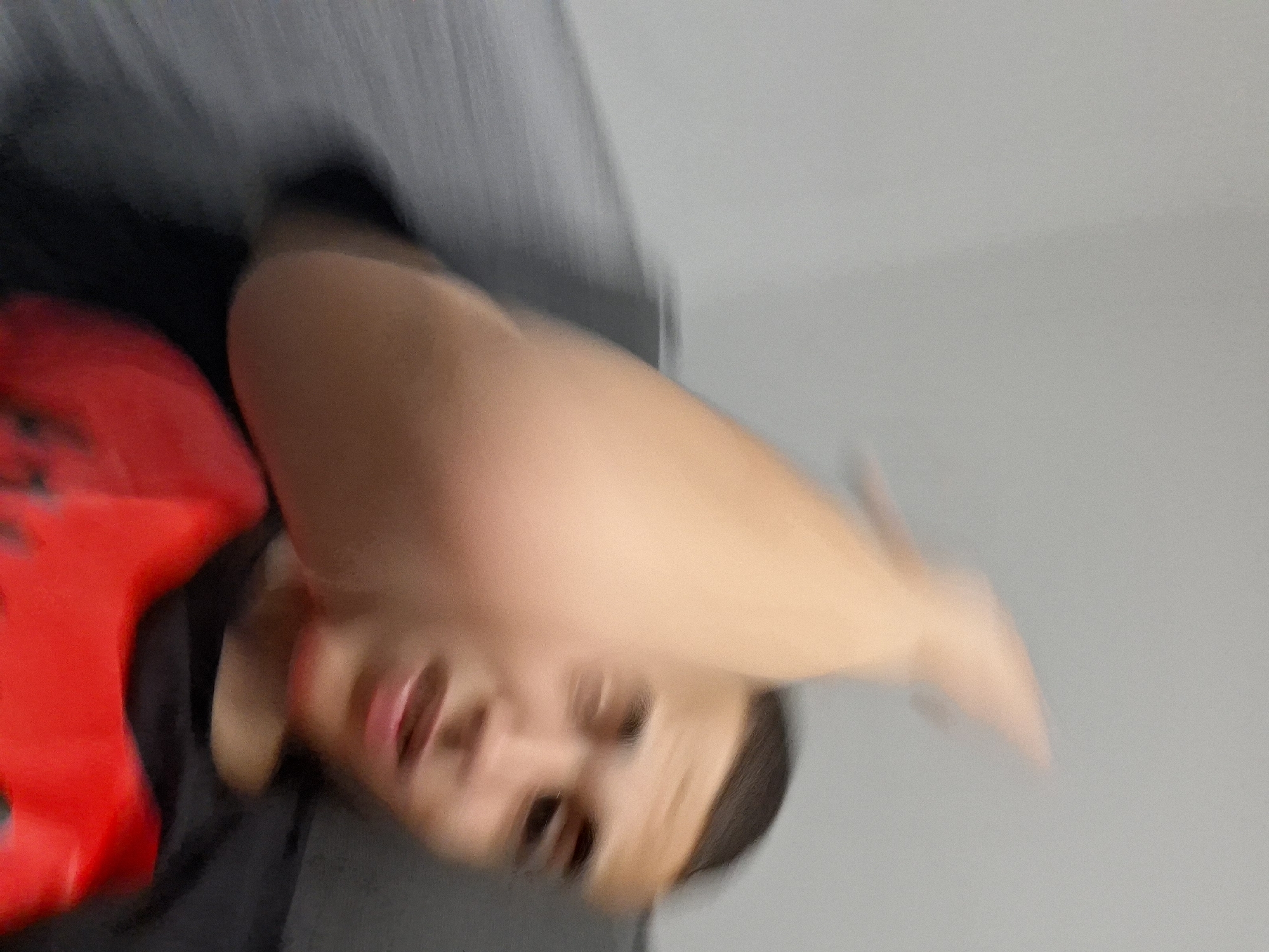 The image depicts a young individual, likely male, with short, dark hair, captured in motion. The subject's right arm is significantly blurred as it is raised and swung forward, occupying a large portion of the foreground. Their face, positioned to the right of the arm, is also motion-blurred, revealing indistinct features and an open mouth. The individual is wearing a black crew-neck shirt over a red garment or object with subtle dark patterns. The background consists of a plain, light gray wall, also blurred, suggesting the subject is seated or leaning against a dark gray, upholstered surface, possibly a sofa. The image does not contain any visual information to determine its geographical location, including the city of Basarbovo, Bulgaria.