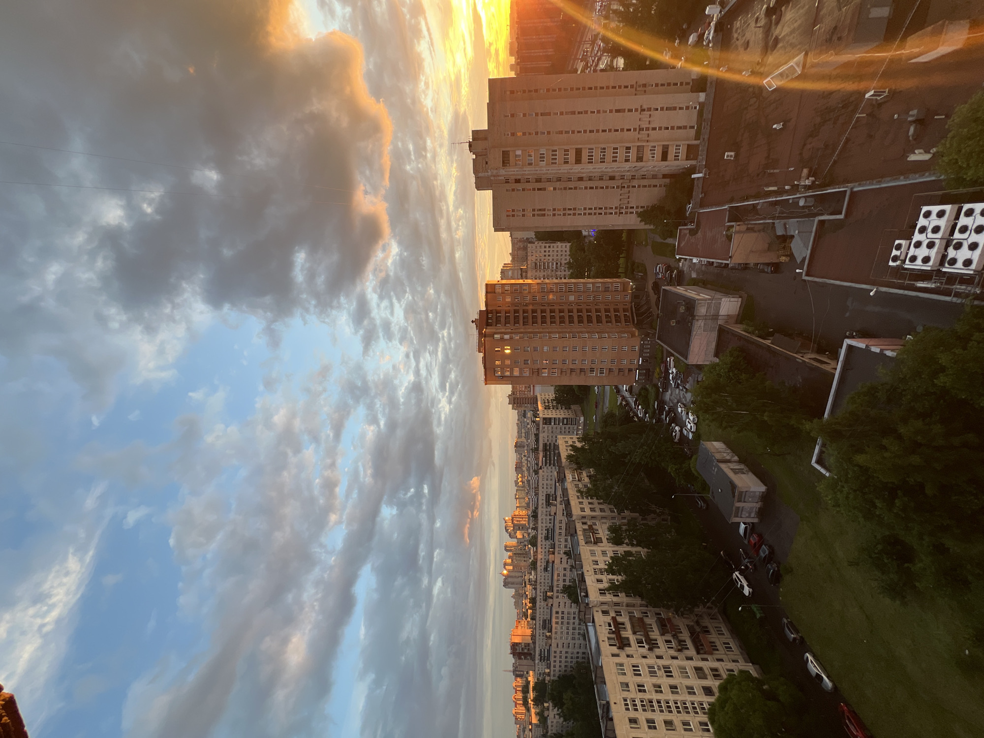 This is a vertical, wide-angle shot of a cityscape at sunset. The sky is filled with dramatic clouds, some tinged with the warm hues of the setting sun, while others are a deep gray. In the foreground, the clouds occupy the left side of the frame, with streaks of blue sky visible. The right side of the image shows a dense urban environment with tall, residential buildings, their facades a light-colored brick. The buildings are a mix of styles, some appearing older with more uniform window patterns, others more modern.
The scene depicts the rooftops of these buildings, some flat with visible air conditioning units, others tiled and brown. A street is visible between the buildings, with parked cars lining it, most appearing to be sedans. Green trees are interspersed throughout the urban landscape, especially along the street and in courtyards. The overall impression is one of a densely populated urban area experiencing the transition from day to night, with the dramatic sky adding an element of natural beauty to the man-made environment. The time of day is clearly sunset, suggested by the warm light illuminating the clouds and the side of the buildings. The weather appears to be partly cloudy. There is no visible text in the image.
