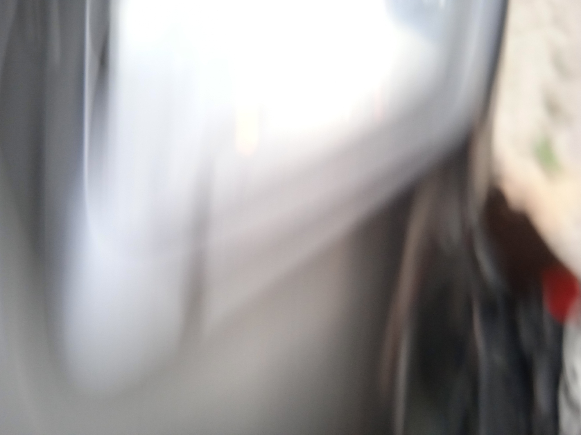 The image is a blurry, abstract photograph taken indoors. The lighting is bright, with large areas of white and grey, suggesting overhead lighting or natural light reflecting off surfaces. On the right side of the image, there is a darker, out-of-focus area that appears to be fabric or clothing, with a hint of red visible. The left side of the image is dominated by a large, smooth, reflective grey surface. Without clear details or context, it is impossible to determine the specific location within Cileunyi, Indonesia, or to identify any specific objects, people, or actions. The image captures a moment with low visual clarity, focusing on light and shadow.