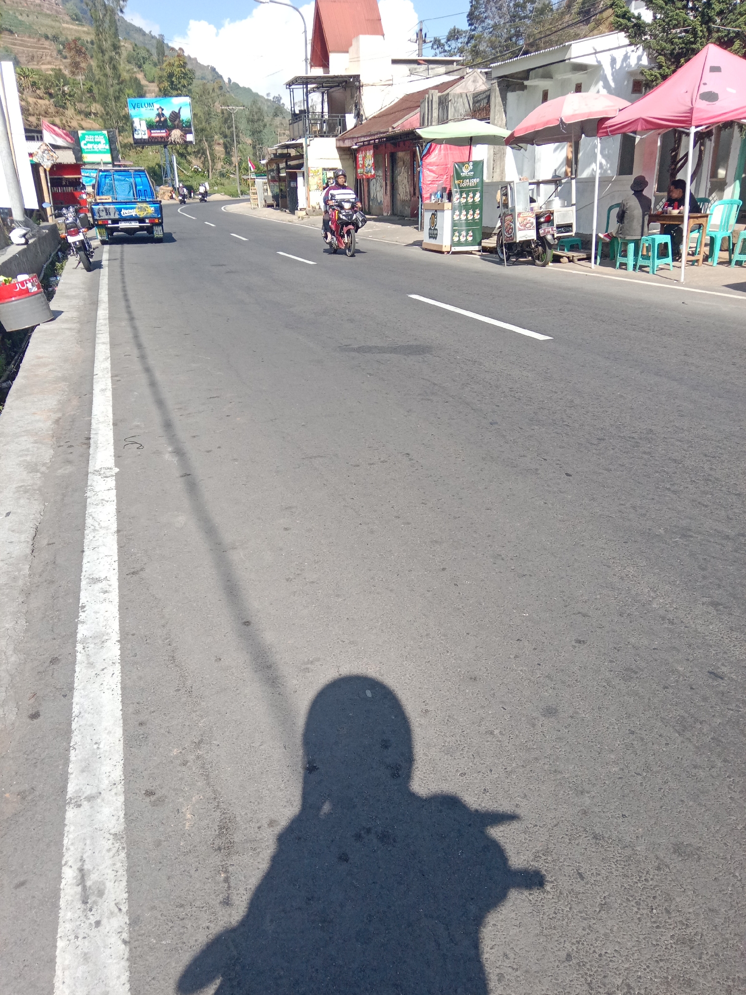 The image depicts a street scene in Parikesit, Indonesia, on a sunny day. A paved road with a white line marking the lane is visible, with a steep hillside covered in trees and buildings on the left. Several vehicles are present, including a blue pickup truck parked on the side of the road and a motorcycle in motion. On the right side of the road, there are several small shops or stalls with awnings, and people are seated at tables. The foreground shows the shadow of the photographer, who appears to be on a motorcycle, with a helmet visible in the shadow. The environment suggests a rural or semi-urban area with commercial activity along a main thoroughfare.