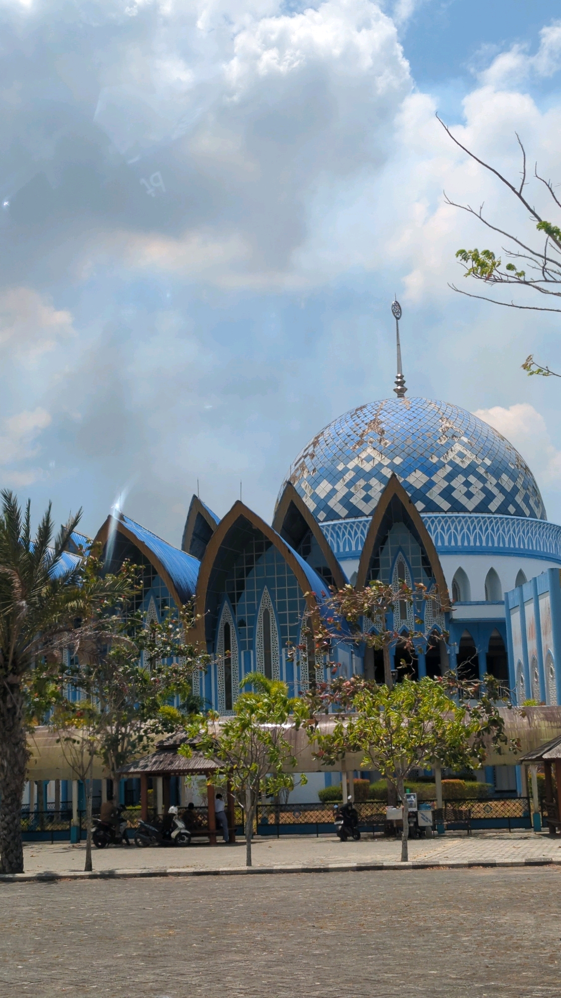 The image shows the exterior of a mosque with a large blue and white patterned dome and multiple smaller arched blue roofs. The mosque is painted in white and blue. In the foreground, there is a paved area, and a row of trees and bushes separates it from the mosque. There are several people and motorcycles near a small open-air pavilion with a thatched roof. The sky is blue with scattered clouds. The image is taken from a low angle, looking up at the mosque. The location is identified as the city of Gunungtinggi, Indonesia.