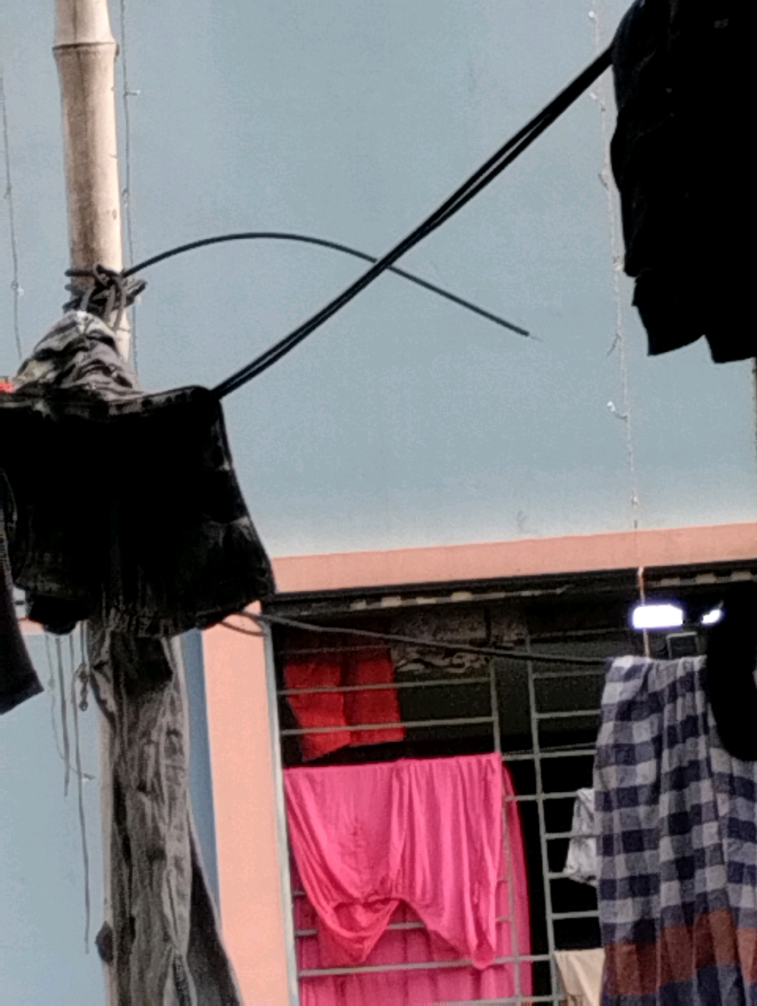 A rooftop laundry scene in Dhaka, Bangladesh, features a bamboo pole on the left, partially obscured by dark clothing hanging on clotheslines. A black garment is prominently displayed, with other dark and grey textiles also visible. Below, a building with a reddish-orange overhang has a window with a metal grate. Through the grate, vibrant pink and red fabrics are draped, suggesting more laundry. A blue and white checkered garment hangs to the right, along with other unidentifiable fabric. The sky in the background is a pale blue. The scene depicts everyday domestic life in a densely populated urban environment.