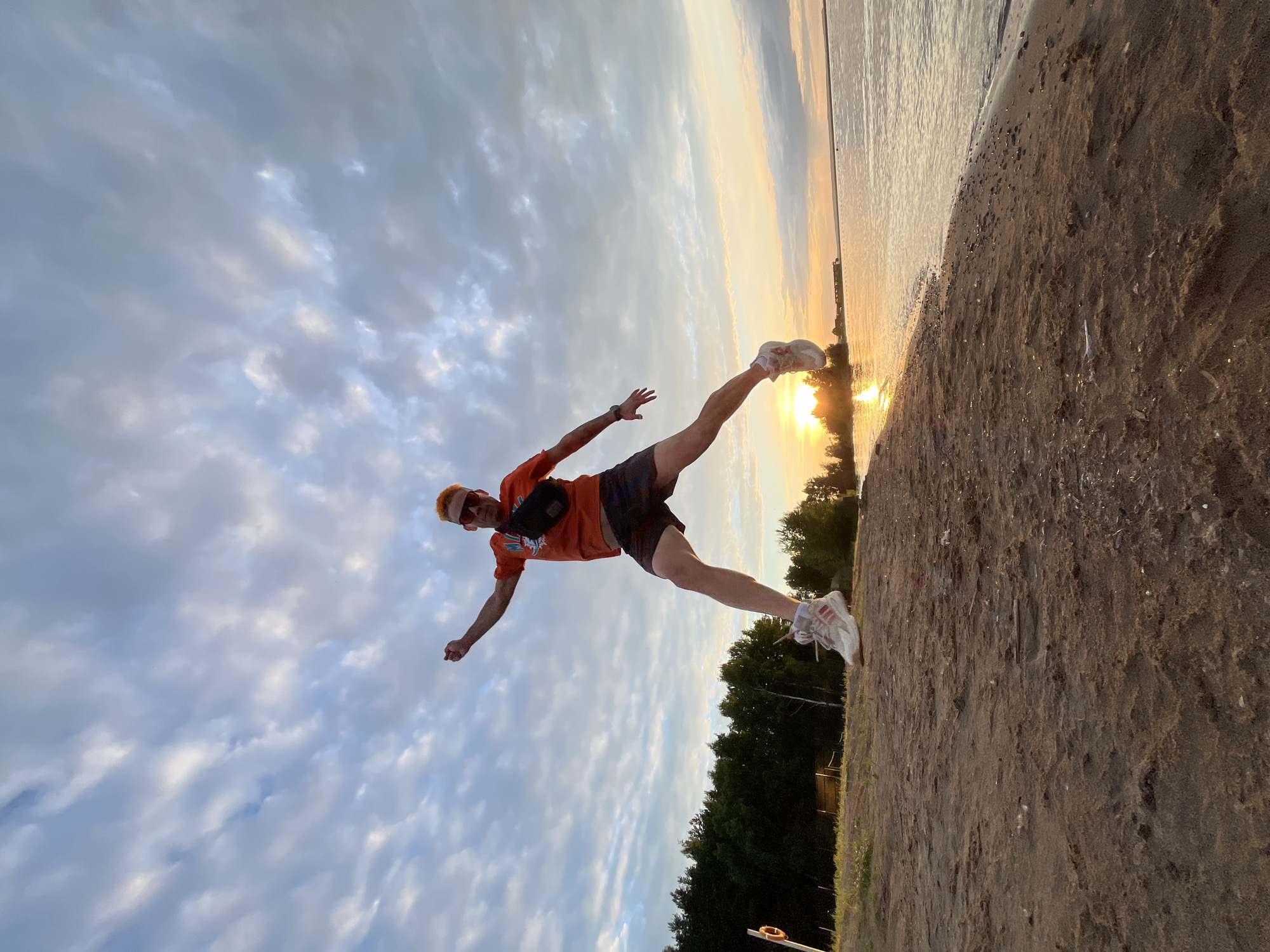 A person is captured mid-air, performing a dynamic jump on a sandy beach next to a body of water. They are wearing an orange t-shirt, dark shorts, a black fanny pack, sunglasses, and white athletic shoes. Their body is angled, with one leg extended upwards and the other bent, and arms outstretched. The sky above is filled with dramatic, wispy clouds, catching the warm, golden light of the setting sun, which is visible near the horizon, reflecting on the calm water. A line of trees borders the beach, and in the distance, structures and a pier suggest a developed area. The overall impression is one of energy and freedom, likely during a warm summer evening. The location is specified as Chaikovsky, Russia.