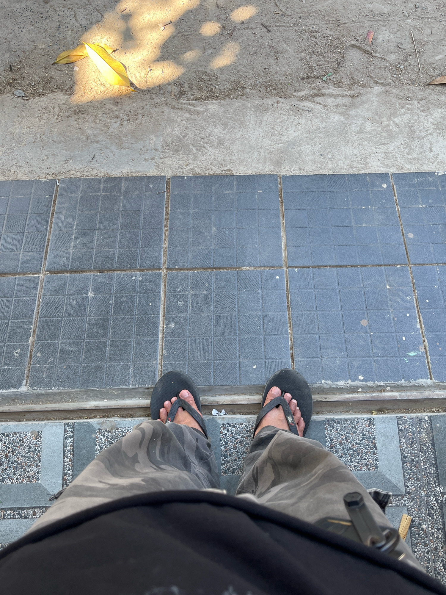 The image is a first-person perspective shot of a person's legs and feet, taken outdoors. The person is wearing black flip-flops and camouflage pants. They are standing on a paved surface that transitions from a textured grey tile to a different patterned grey tile. In the background, a rough concrete area with scattered debris and fallen leaves is visible, partially illuminated by sunlight creating shadows. The scene appears to be a sidewalk or walkway in a public area. The location in Tungkaran Pangeran, Indonesia, is inferred from the prompt but not visually evident in the image itself.