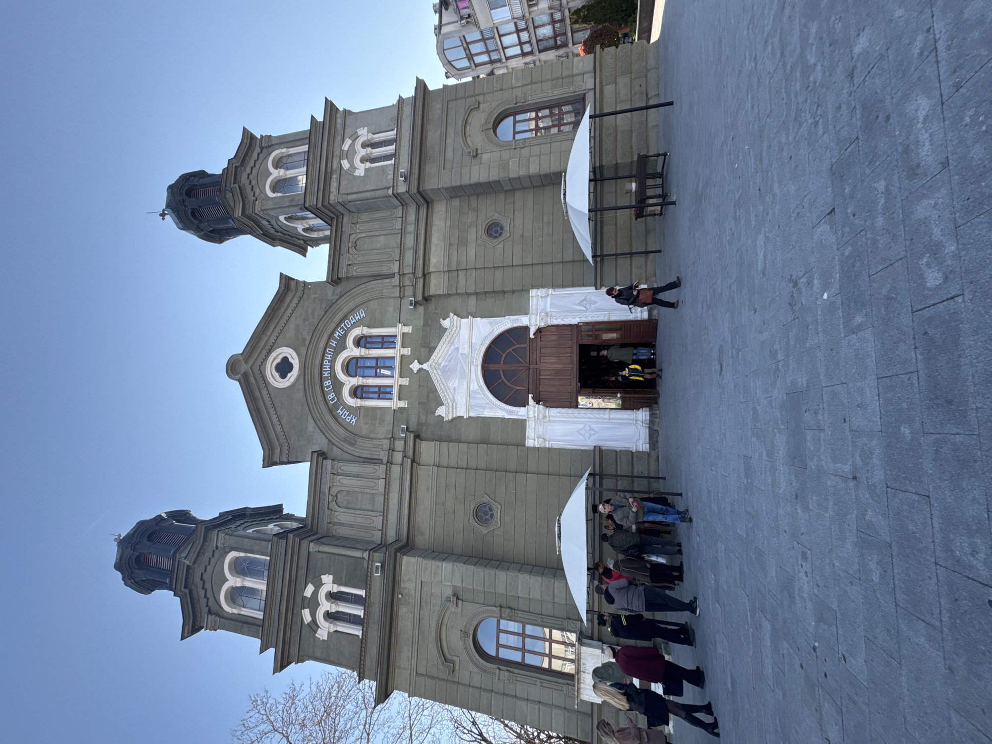 The image shows the Cathedral of Saints Cyril and Methodius, located in the city of Burgas, Bulgaria. The cathedral is an impressive stone structure with two prominent towers. A group of people are standing near the entrance. A white canopy provides shade near the entrance. The foreground shows a paved area. The sky is clear blue.
