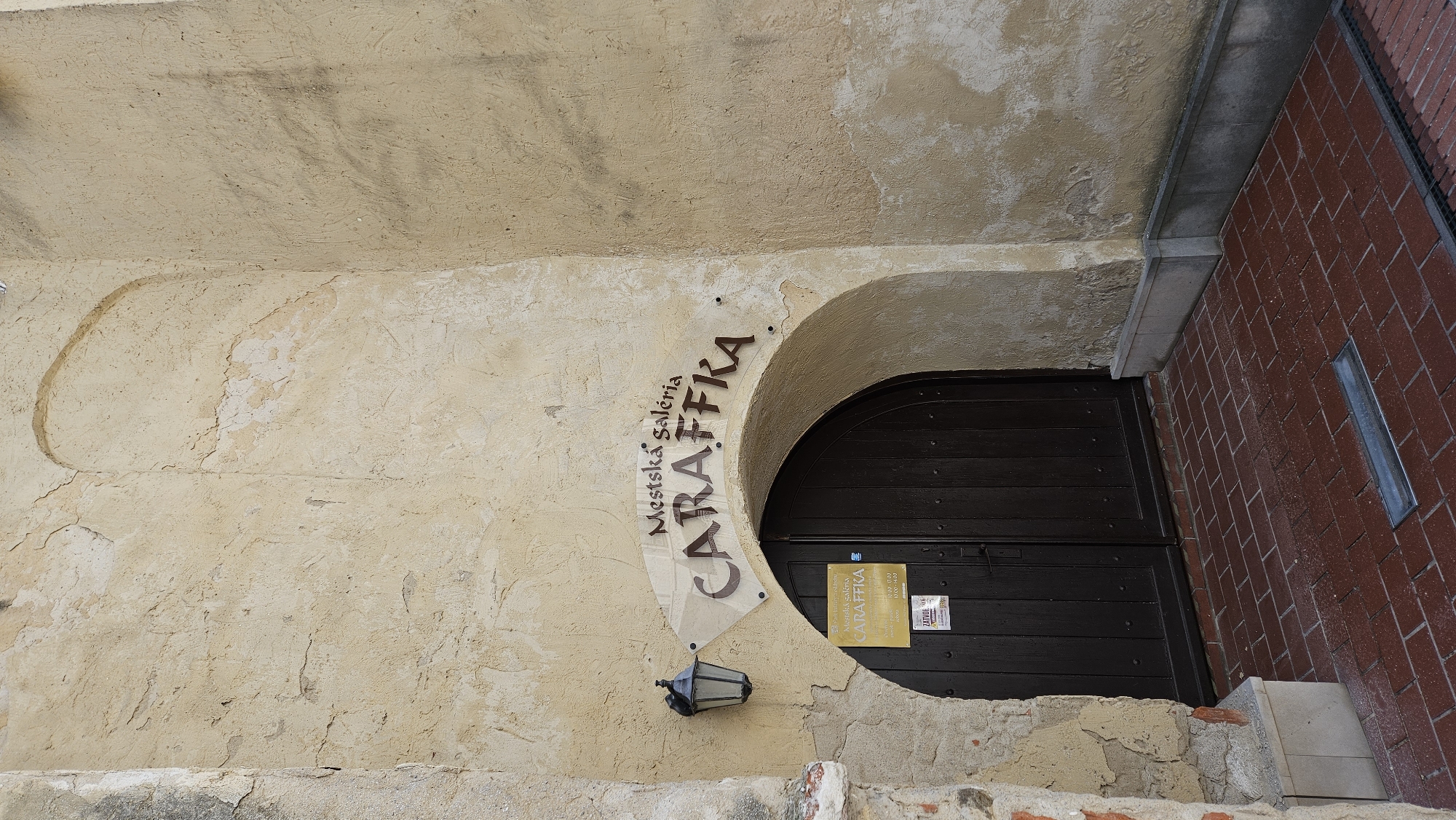 The image shows the entrance to the Caraffka Municipal Gallery in Prešov, Slovakia. The entrance features a large, arched wooden door set into a weathered, yellow stucco wall. Above the door, a sign with the text "Mestská galéria CARAFFKA" is mounted on a light-colored, curved plaque. To the left of the sign, an old-fashioned black lantern is attached to the wall. Below the main sign, a smaller sign is affixed to the door, displaying gallery hours and information. The ground in front of the entrance is paved with red brick. The overall scene depicts an outdoor architectural facade with clear signage indicating a cultural institution.