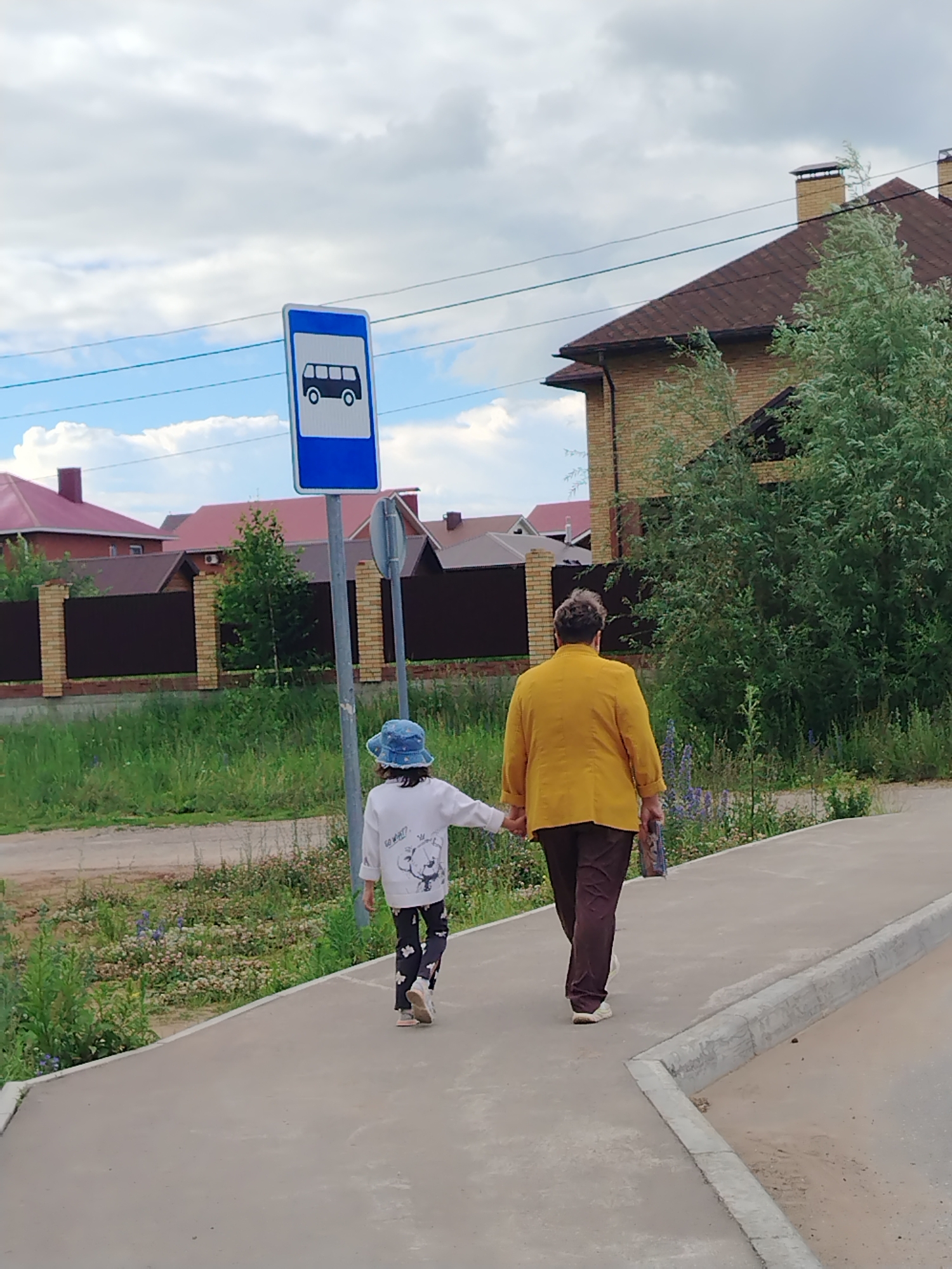 The image shows a person and a child walking hand-in-hand on a sidewalk in Noviy, Russia. A bus stop sign is visible on the left side of the image. The background features houses with red roofs and green trees, suggesting a suburban or semi-rural setting. The sky is partly cloudy. The person is wearing a yellow jacket and dark pants, while the child is wearing a white sweatshirt, a blue hat, and patterned pants. They are walking away from the camera towards the right side of the frame.