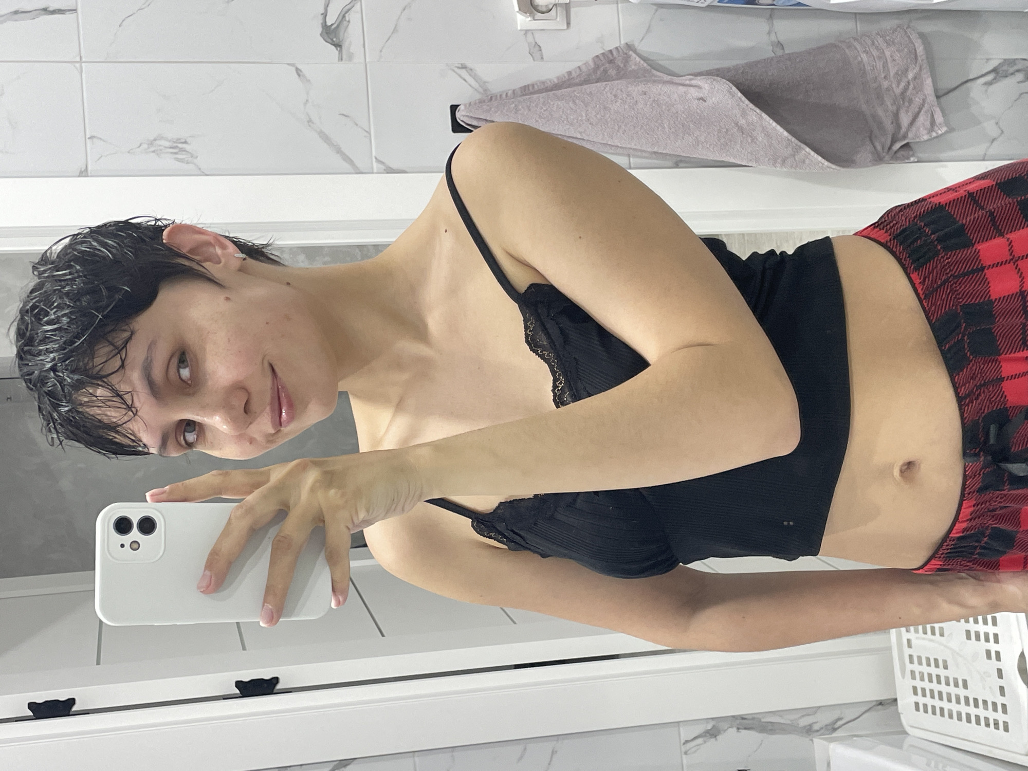 A person with wet, short dark hair is taking a selfie in what appears to be a bathroom. They are wearing a black tank top with lace trim and red and black plaid pajama bottoms. The person is holding a white smartphone in their right hand. The bathroom features white marble-patterned tiles, a white door or window frame, and a grey towel hanging on the wall. The scene lacks sufficient detail to confirm its location in Izhevsk, Russia.