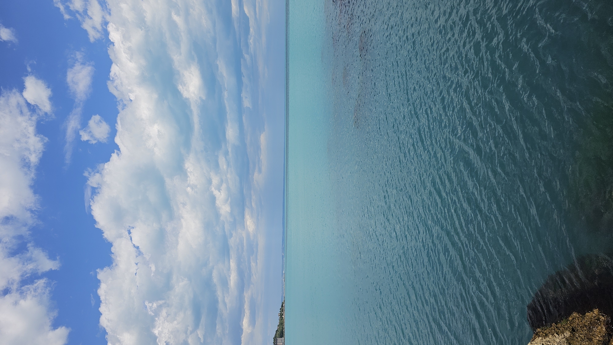 The image depicts a body of water meeting the sky, possibly at a beach in Varna, Bulgaria. The left side of the image shows a vibrant blue sky with puffy white clouds. The right side shows water of varying blue hues, with darker areas suggesting deeper water or shadows. A rocky landmass is visible in the lower right corner. The coastline is faintly visible as a thin, dark line along the horizon near the center of the image.