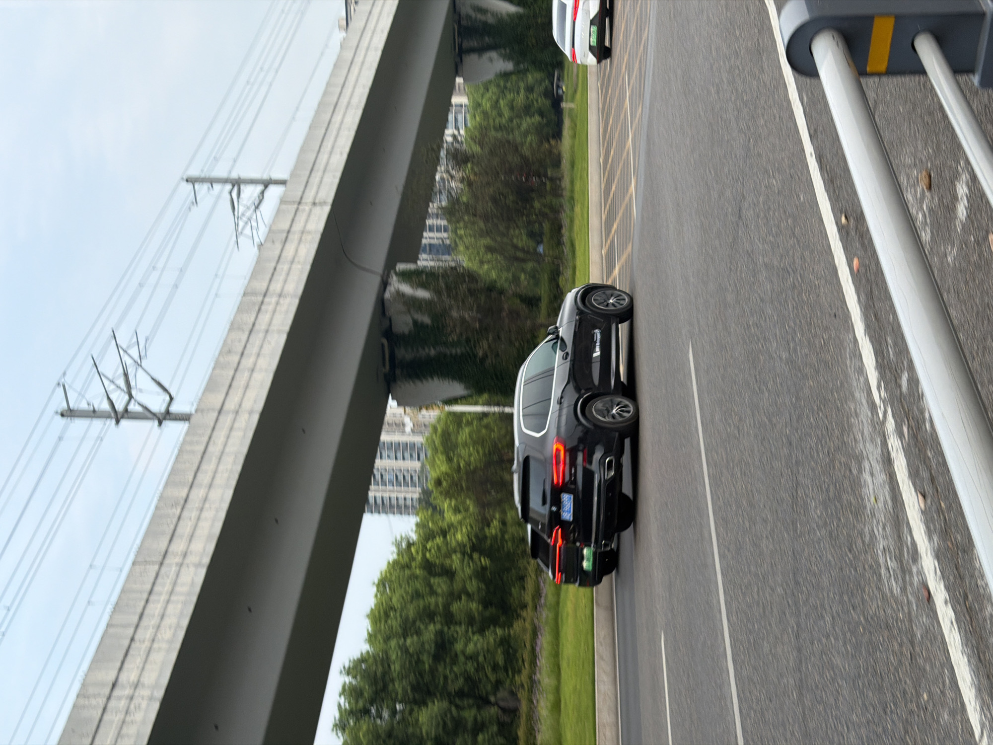 The image shows a road with vehicles under an elevated structure, possibly a highway or overpass. A black SUV, possibly a BMW, is in the foreground, moving on the right side of the road. Another dark-colored SUV is behind it. A white vehicle is visible further ahead. The environment appears to be a city with trees and buildings in the background. The sky is overcast. The elevated structure has electrical wires running along it. The scene is likely captured in Jiangjin, China, given the presence of Chinese license plates and the architectural style.