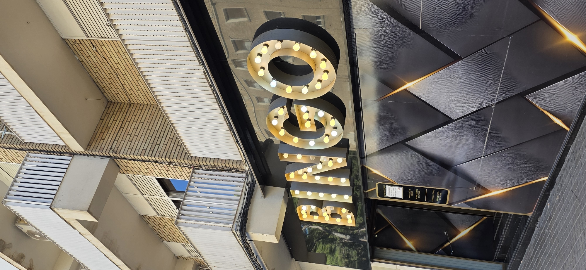 The image shows a close-up, low-angle view of the exterior of a building with a prominent sign. The sign is composed of large, illuminated letters spelling out "BINGO" with vintage-style light bulbs. The letters are dark grey and three-dimensional, with the bulbs casting a warm, yellow glow. The sign is mounted on a reflective, black surface, which mirrors the illuminated letters and the surrounding environment. To the left of the sign, the building's facade is visible, featuring a combination of light-colored louvers and textured, yellowish bricks. The overall scene appears to be an outdoor setting, likely in a commercial or entertainment district. The lighting suggests it might be daytime, possibly late afternoon or early evening, given the warm glow of the lights. No people are visible in the frame. There is text on a dark rectangular sign below the "BINGO" sign, but it is too blurry to read. The architectural style of the building, with its distinct brickwork and louvers, could be a clue to the location. Given the context of Huesca, Spain, this might be the facade of a bingo hall or a similar establishment. The reflective surface of the building and the prominent signage contribute to a modern and eye-catching aesthetic.