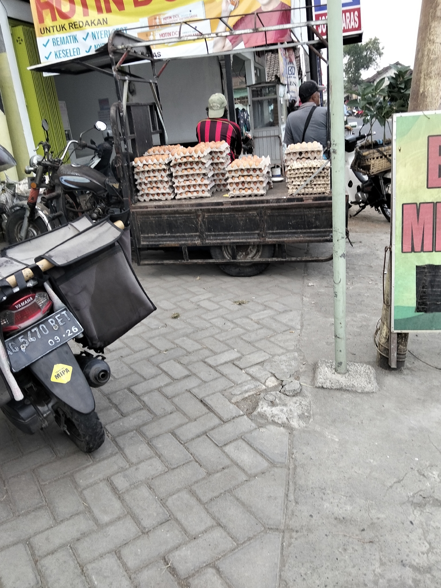 This is a photograph of a street scene in Gempolan, Indonesia. In the foreground, a black motorcycle with a red license plate that reads "AG 5470 BET 09-26" is parked on a paved surface. The motorcycle has a black bag attached to the back. Behind the motorcycle, a black truck with stacks of egg cartons is visible. Two men are standing near the truck, one wearing a black hat and a grey shirt, and the other wearing a striped shirt. In the background, there are buildings and trees. A green pole with a sign is also visible on the right side of the image. The overall scene depicts a bustling street market or roadside stall.