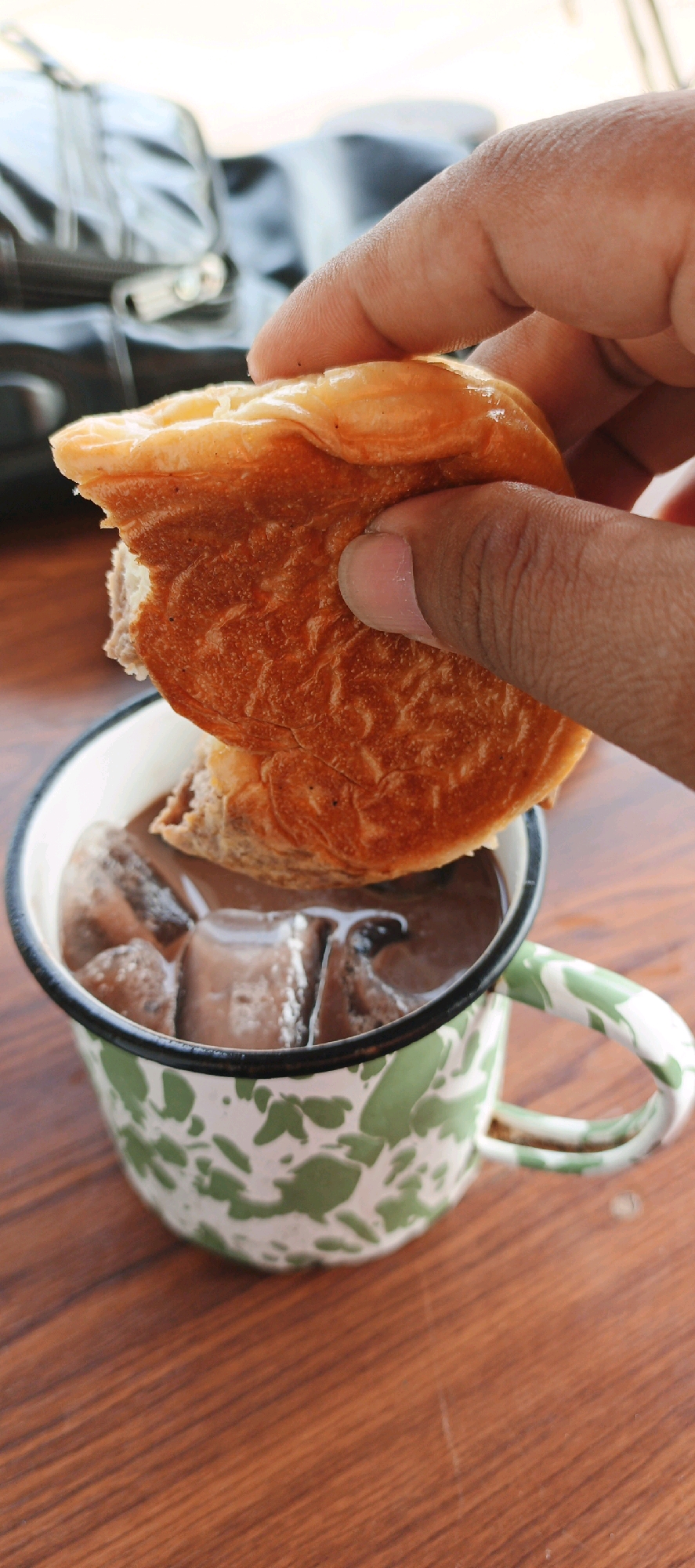A person's hand is holding a piece of a pastry over a mug of iced chocolate. The mug is white with a green abstract pattern and has a green rim and handle. The pastry is golden brown and appears flaky. The chocolate drink is dark brown and filled with ice cubes. The scene is set on a wooden table. In the background, a blurred dark bag is visible. The image appears to be taken indoors or in a shaded outdoor area. This scene could plausibly be found in a cafe or eatery in Yogyakarta, Indonesia, a city known for its rich culinary scene and cultural offerings.