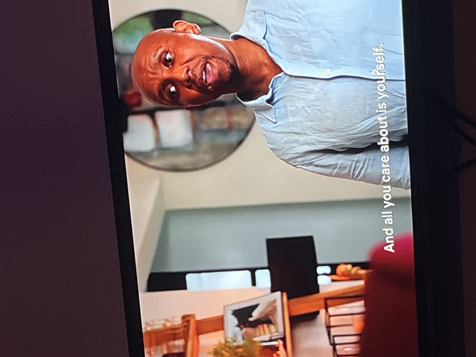 The media file is a photograph taken indoors, likely in a home or studio. The primary subject is a Black man, bald with a goatee, wearing a light blue collared shirt. He is looking directly at the camera with an open-mouthed expression, conveying surprise or excitement. His head is tilted to the left.
Behind the man, a circular mirror reflects a blurred image of what appears to be shelves filled with books or decorative items. The background also shows parts of a room with furniture, including chairs and a table, suggesting a domestic setting. A framed picture is visible on the table.
On the right side of the image, vertical white text is superimposed over the man's shirt and the background. The text reads, "And all you care about is yourself." The lighting suggests it is daytime, with no indication of specific weather conditions. The overall emotion conveyed by the man's expression is one of heightened emotion or a moment of surprise. The scene appears to be staged or captured during a recording session.