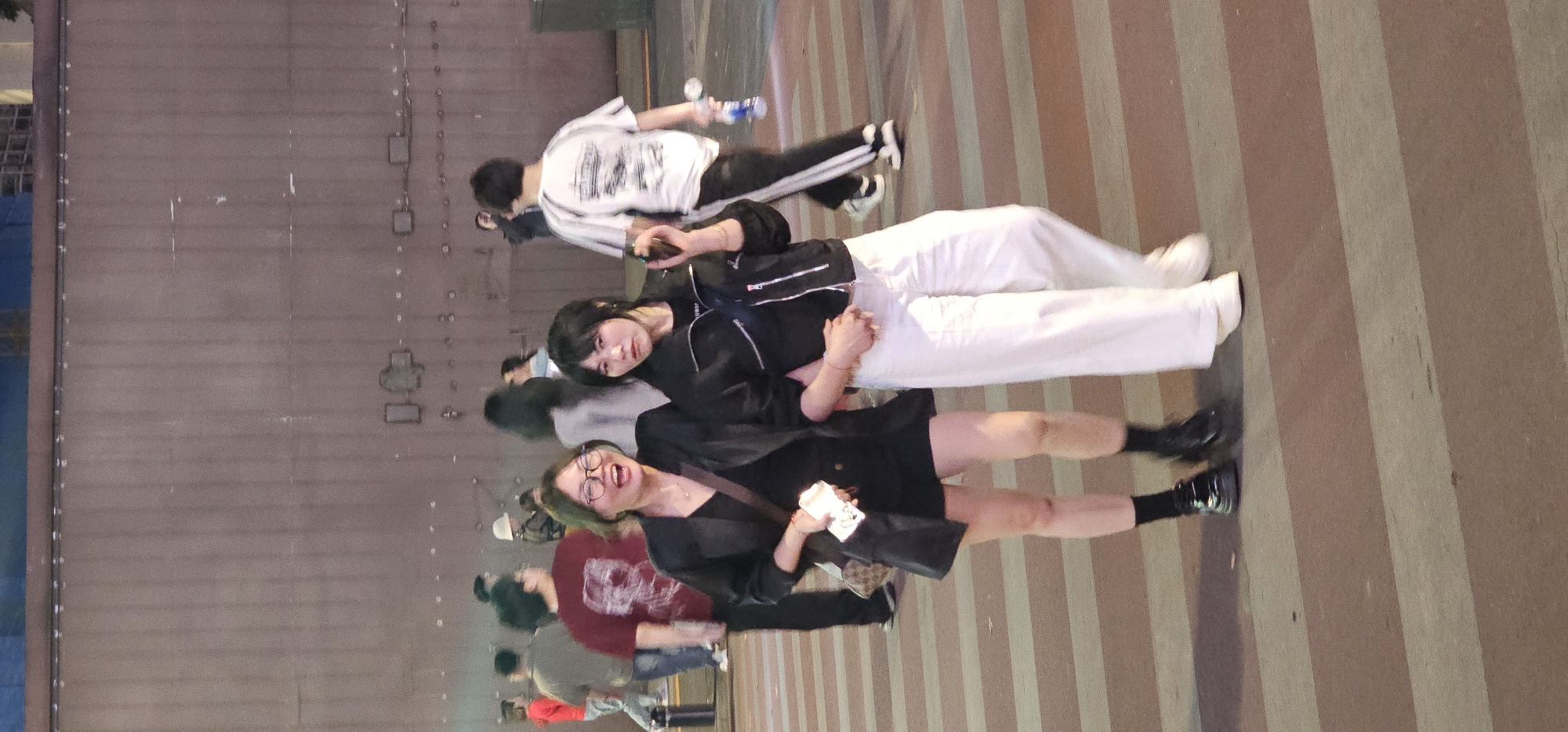 The image captures a street scene in Yuzhong District, China. The foreground features two women standing close to each other, apparently posing for a photo. The woman on the left is smiling broadly with her mouth open, wearing glasses, a black dress, and black boots with black socks. She holds a white object in her right hand, possibly a paper cup or takeout container, and has a black purse slung over her left shoulder. The woman on the right has straight black hair and is wearing a black jacket with a small backpack and white pants, holding the other woman's hand with hers. Behind them are other people and objects, including a man wearing a white shirt, and what appears to be a wall with metallic features. The ground has brown stripes, suggesting a crosswalk or patterned walkway. The lighting suggests it is nighttime.