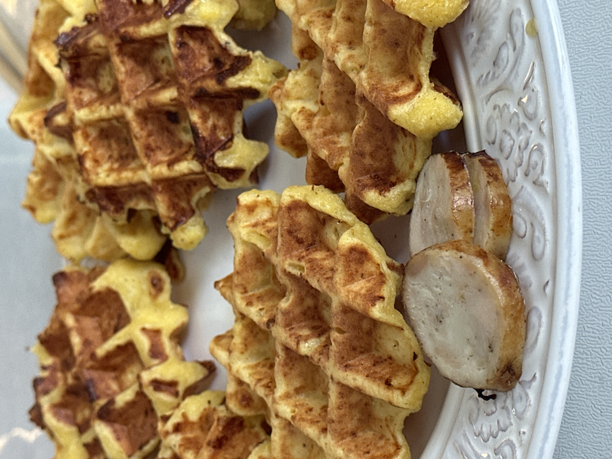 The image displays a close-up view of several golden-brown waffles, some partially visible, arranged on a white plate with embossed decorative edges. Two slices of cooked sausage are positioned in the foreground on the left side of the plate, below the waffles. The lighting suggests an indoor setting. There are no visible people or signs of a specific urban environment, thus the location in the city of Рабочий, Russia cannot be determined from the image alone.