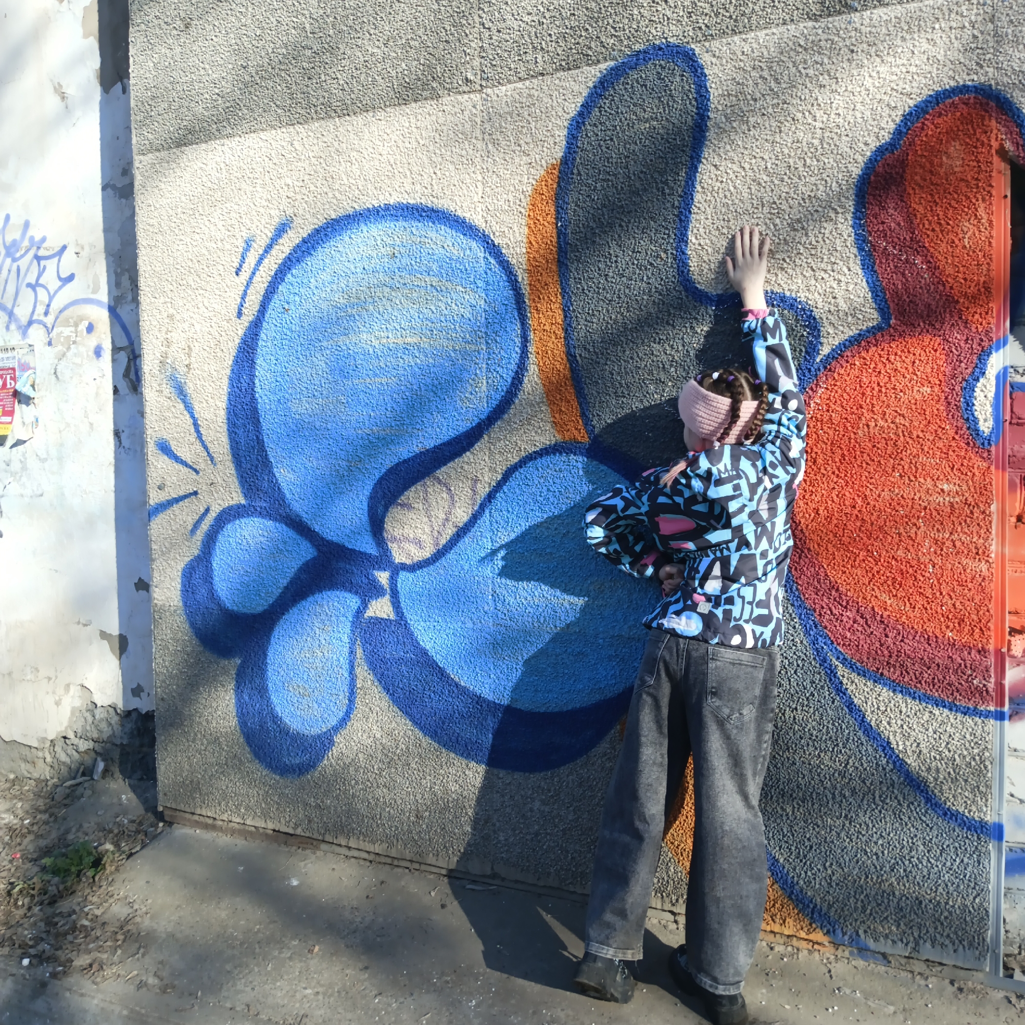 A person wearing a patterned jacket and jeans stands against a concrete wall covered in graffiti. The graffiti consists of stylized shapes in blue, orange, and red. The person has a light pink headband and dark braided hair. Their right arm is extended, with their hand touching the wall. The background includes a section of deteriorating white wall and a paved area with debris. The location is the city of Chaikovsky, Russia.
