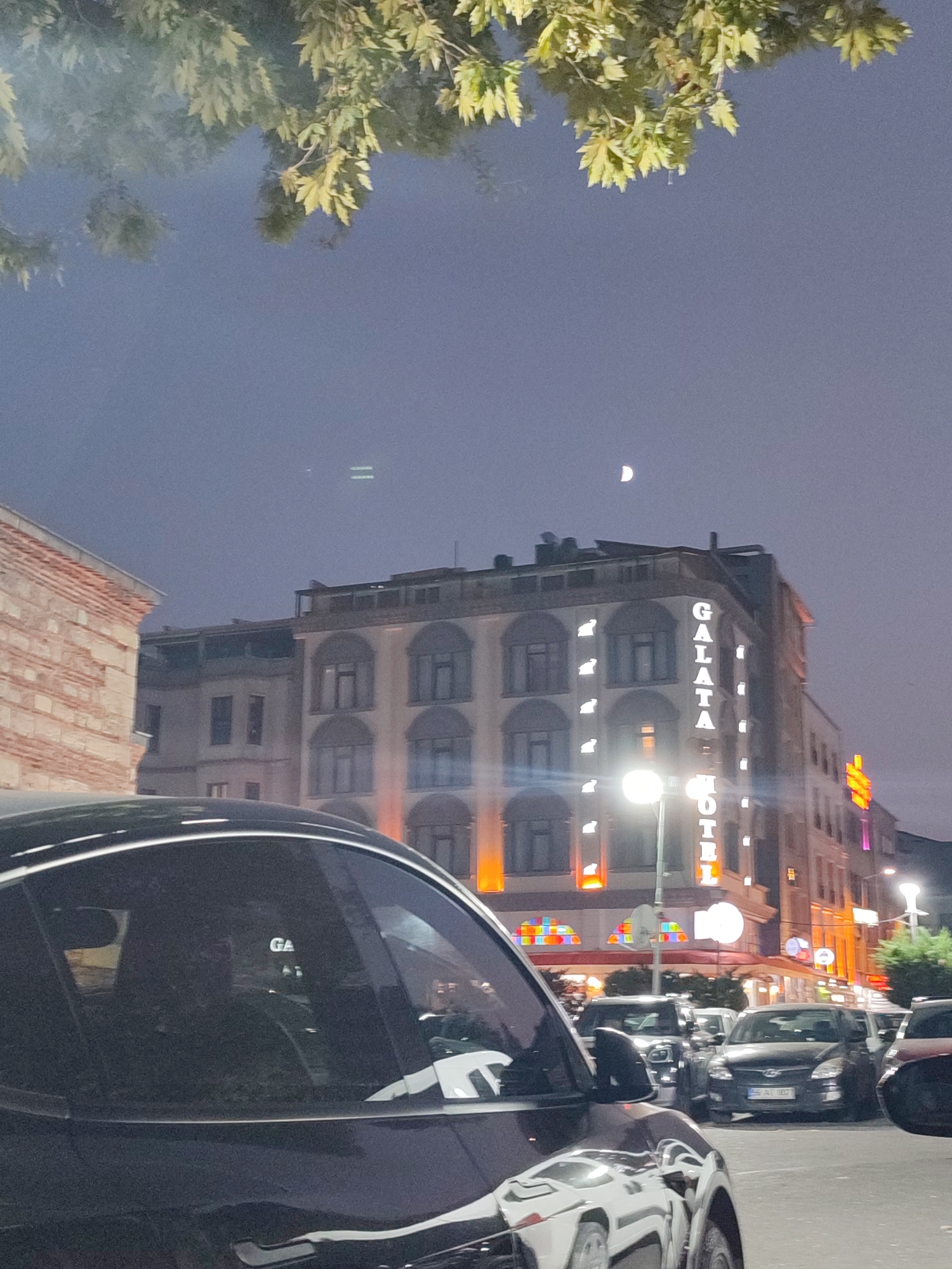 A street view in Beyoğlu, Turkey, at dusk. A dark-colored car is in the foreground, partially obscuring a row of parked cars behind it. In the background, the "GALATA HOTEL" is illuminated with neon lights. The sky is hazy, with a sliver of moon visible. Trees with green and yellow leaves hang over the scene from the top.