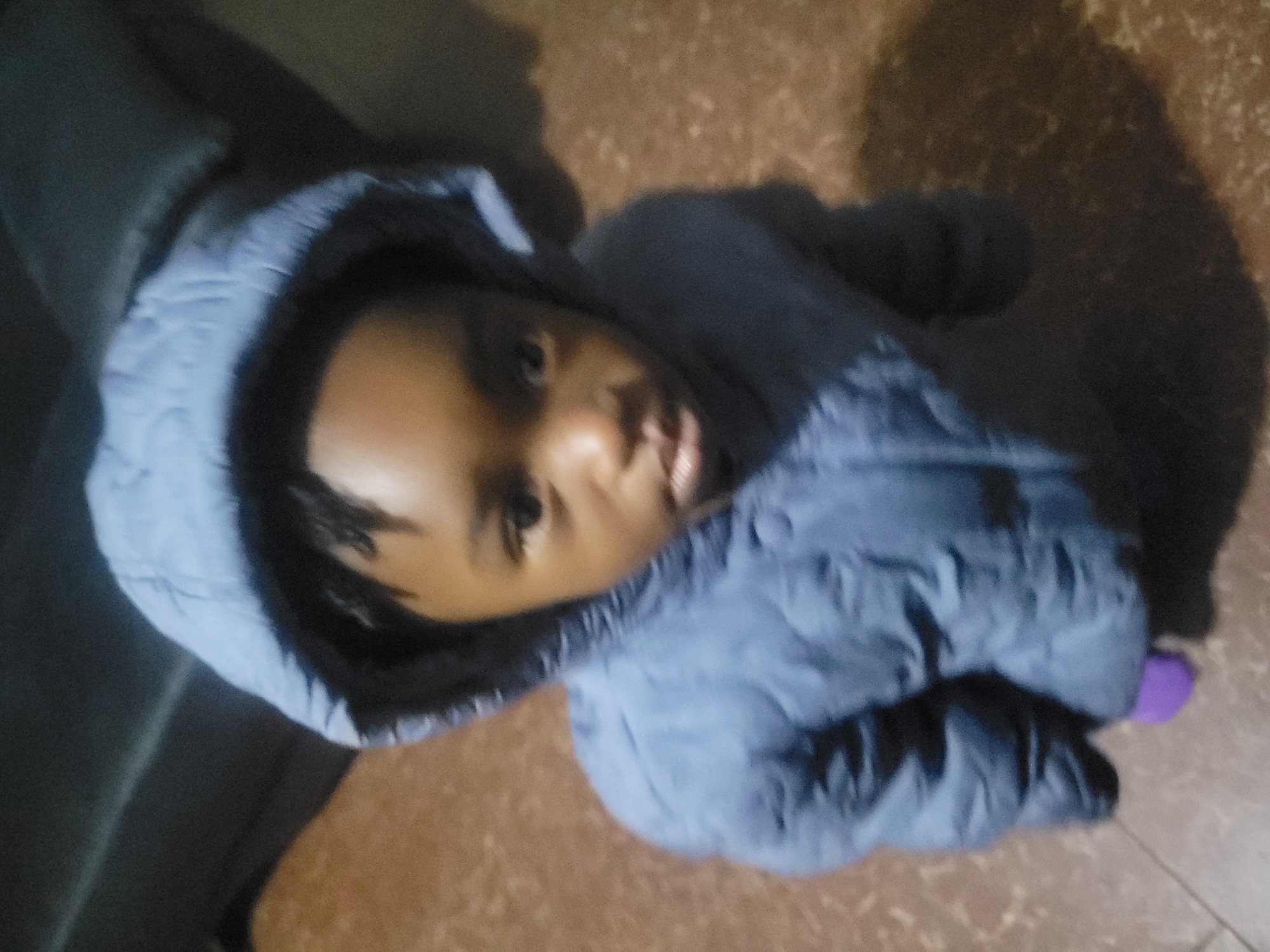 A child wearing a hooded, blue puffy jacket looks up at the camera. They are lying on a floor with a mottled brown pattern. The jacket hood is lined with black material. A purple object is partially visible near the bottom right corner of the image. A blurry dark object is visible in the upper left corner. The location is unspecified.