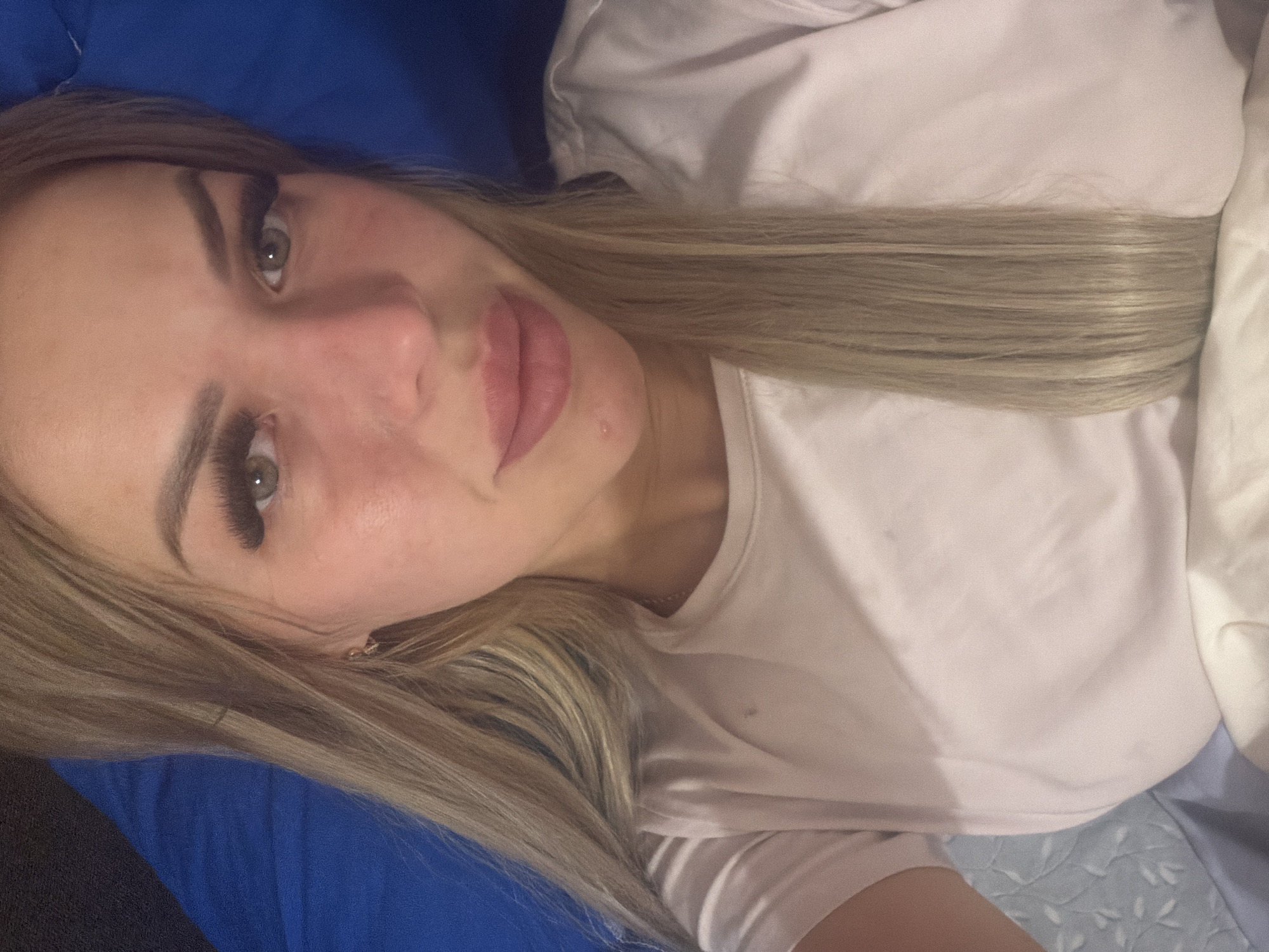 A close-up, overhead, selfie-style shot shows a fair-skinned woman with blonde hair, wearing a light pink t-shirt. She has long, dark eyelashes, light green eyes, and full lips. A small red mark is visible on her chin. She is lying down, with her head resting on a blue pillow. The background is a dark blue surface. The image was taken indoors, likely in a bedroom in Kyiv, Ukraine.