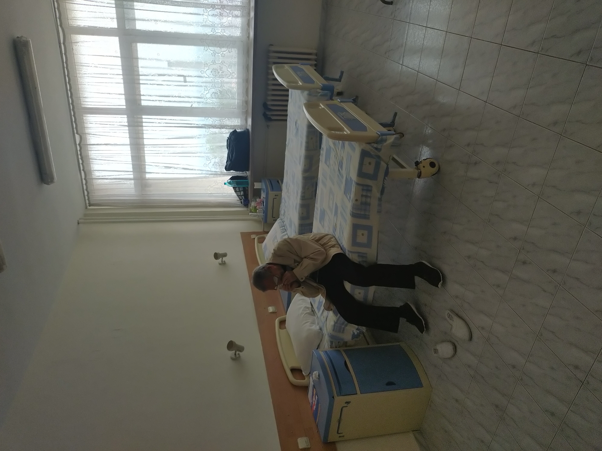The image, rotated 90 degrees counter-clockwise, captures an indoor scene depicting a room in what appears to be a hospital or care facility, likely located in Stara Zagora, Bulgaria. The room is brightly lit by natural light streaming through a large window on the far wall, suggesting it is daytime.
The primary subject is an elderly man, dressed in a light-colored jacket and dark trousers, leaning against one of two beds in the room. His posture suggests he might be resting or experiencing discomfort, and he is holding a small object, possibly a tissue, near his mouth. A pair of white slippers are visible on the light-colored tiled floor near his feet.
The room features two beds with light-colored headboards and footboards, each covered with a blue and white patterned bedspread. Between the beds, a light blue and beige nightstand is present, holding several small bottles and items. A second nightstand is partially visible. A black bag and more bottles are placed on the windowsill. The large window is covered by sheer white curtains featuring a floral pattern, and a radiator is positioned beneath it. The walls are plain white, equipped with two wall-mounted light fixtures. A yellow football can be seen tucked under the second bed. No visible text is discernible in the image.