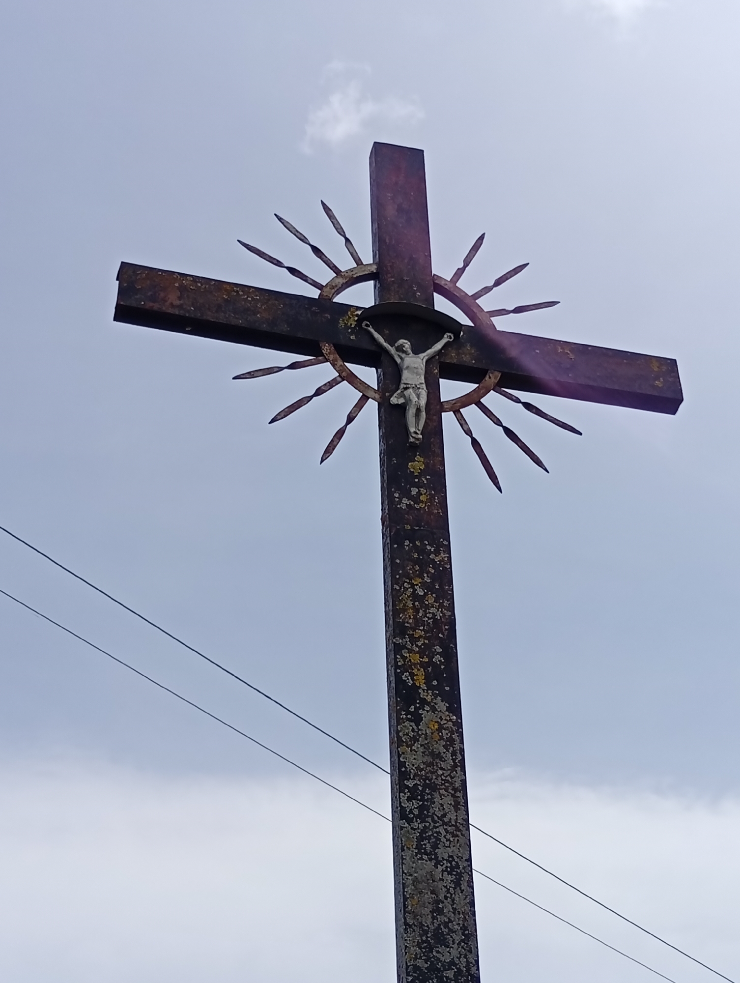 A low-angle, full shot of a weathered, dark metal cross stands against an overcast sky. The cross features a circular halo behind the figure of Jesus Christ. The halo has sharp, pointed rays extending outwards. Jesus is depicted with his arms outstretched and his head bowed, wearing a loincloth. The vertical post of the cross is covered in patches of light green and grey lichen, indicating age and exposure. Two thin, dark wires run diagonally across the lower portion of the image, obscuring parts of the sky and the base of the cross. The scene is devoid of people and any other discernible structures. The location is identified as Krikliai, Lithuania.