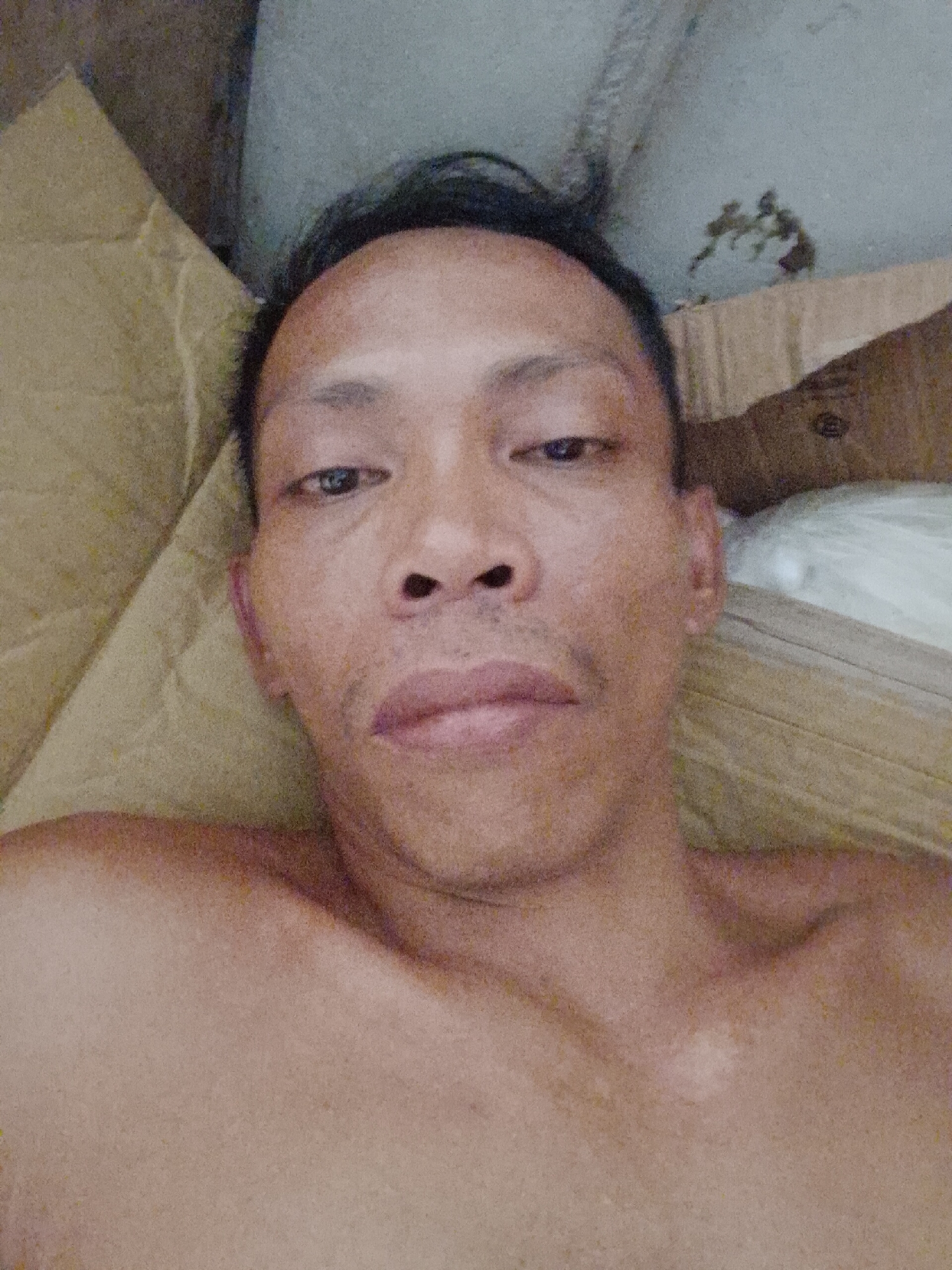 A close-up selfie of a shirtless adult male with short black hair. He is lying down, with a tan-colored surface (possibly fabric or cardboard) behind his head and shoulder. The background also includes a white object and corrugated cardboard. The photo's location is likely Bukit Ketok, Indonesia.