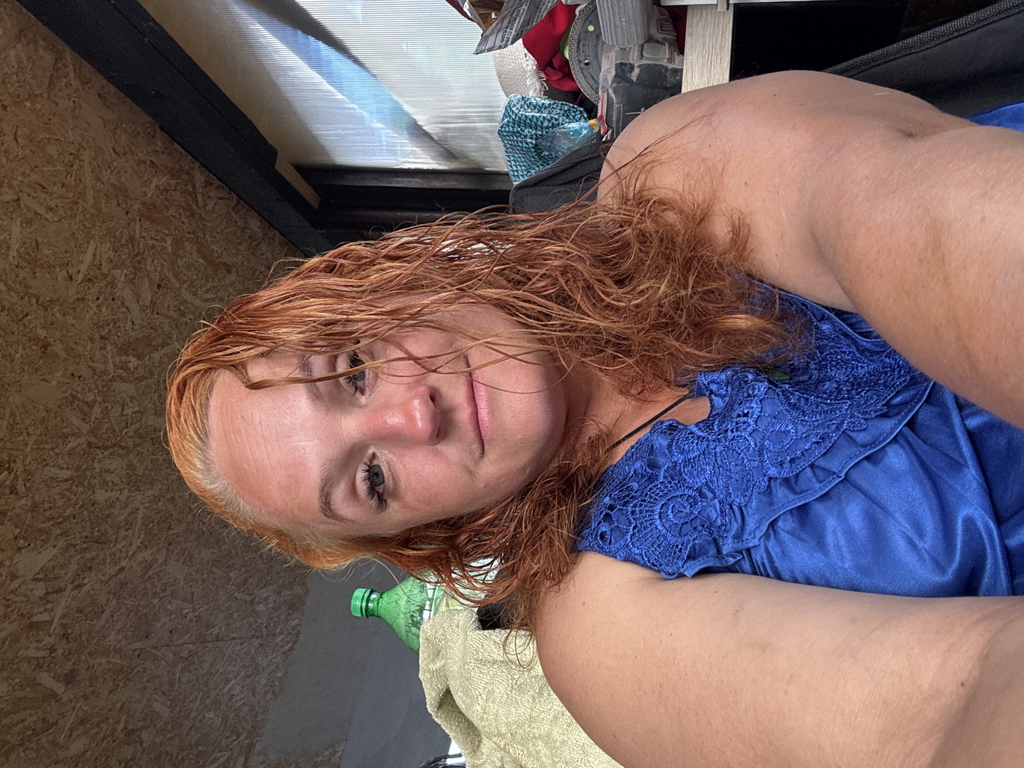 A middle-aged woman with red hair is sitting indoors, looking at the camera. She is wearing a blue dress with lace detail. Behind her, there is a green bottle, a towel, and various objects on a shelf. The walls appear to be made of wood panels. The lighting suggests it is daytime. The location is Chaikovsky, Russia.