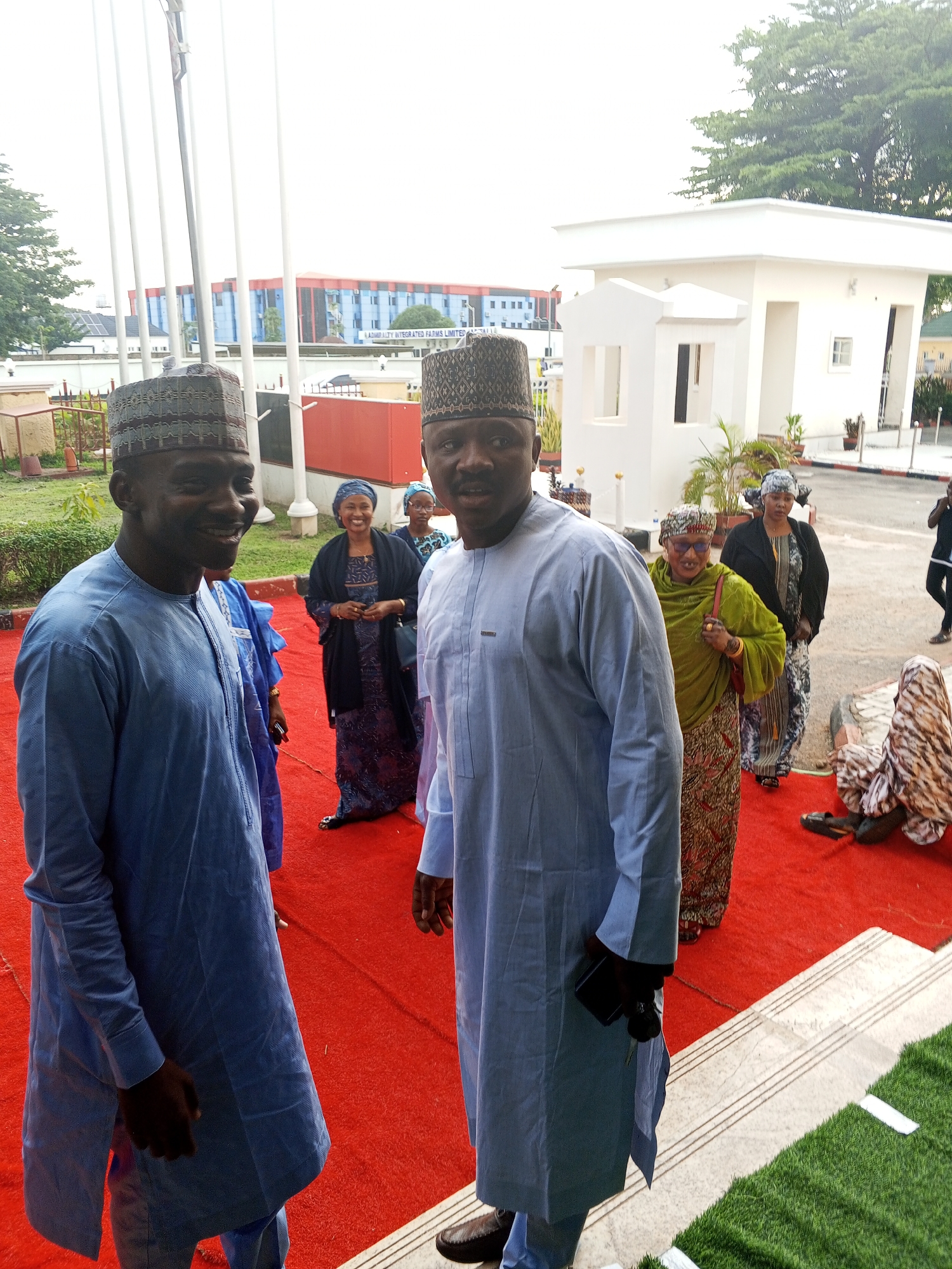 In Abuja, Nigeria, two men in traditional attire stand on a red carpet. The man on the left, wearing a light blue tunic and cap, smiles at the camera. The man on the right, also in a light blue tunic and cap, holds a phone in his hand and looks forward. Behind them, other individuals in similar clothing are visible, some smiling and talking. The background features a modern white building with large windows and architectural details, likely part of an official or institutional complex. Lush green grass borders the carpeted area. The scene suggests a formal gathering or event taking place outdoors in a well-maintained urban environment.