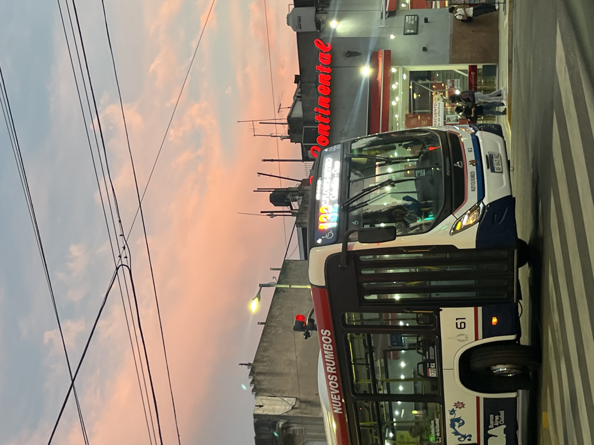 The image captures a street scene in Buenos Aires, Argentina, at dusk, marked by a sky with pink and blue hues. A bus, identifiable by the number "61" and the phrase "NUEVOS RUMBOS" on its side, occupies the foreground. The bus's license plate is "AF 041 NZ."
In the background is a building with the word "Continental" illuminated in red. Pedestrians are present near the building, suggesting commercial activity. Overhead, power lines are visible against the sky. The presence of zebra stripes suggests a pedestrian crossing. A lamppost illuminates the street. The bus also shows some emblems including "Colectivos de la Ciudad", "132 61" and "Buenos Aires Ciudad".