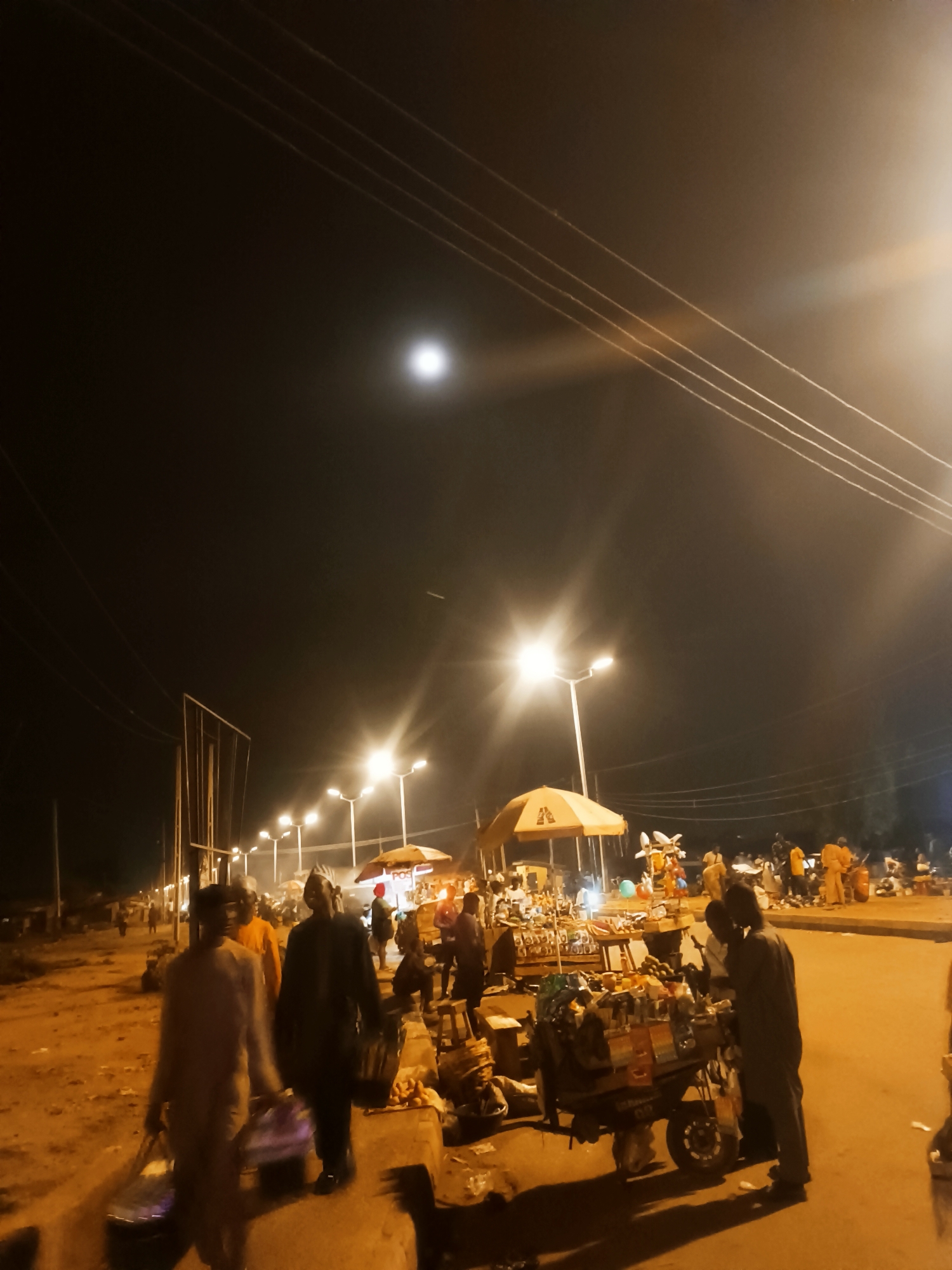 The image depicts a street scene at night in Bukuru, Nigeria. The sky is dark with a visible moon and power lines overhead. The street is illuminated by bright streetlights, with vendors setting up under umbrellas. People are walking and interacting with the vendors, some carrying bags. A cart laden with goods is visible in the foreground. The road surface appears rough and uneven.