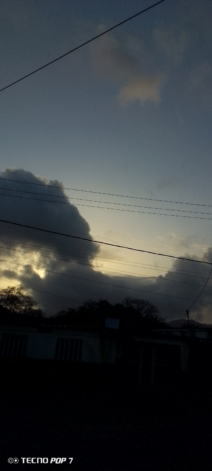 The image shows a cloudy sky at dusk or dawn, with a few visible clouds and some clear sky. Power lines run across the frame. Below, silhouetted trees and buildings are discernible against the fading light. The location is potentially Altagracia, Venezuela, but there are no specific details to confirm this. The scene is dimly lit, making details difficult to discern. A "Tecno Pop 7" watermark appears in the lower left corner.