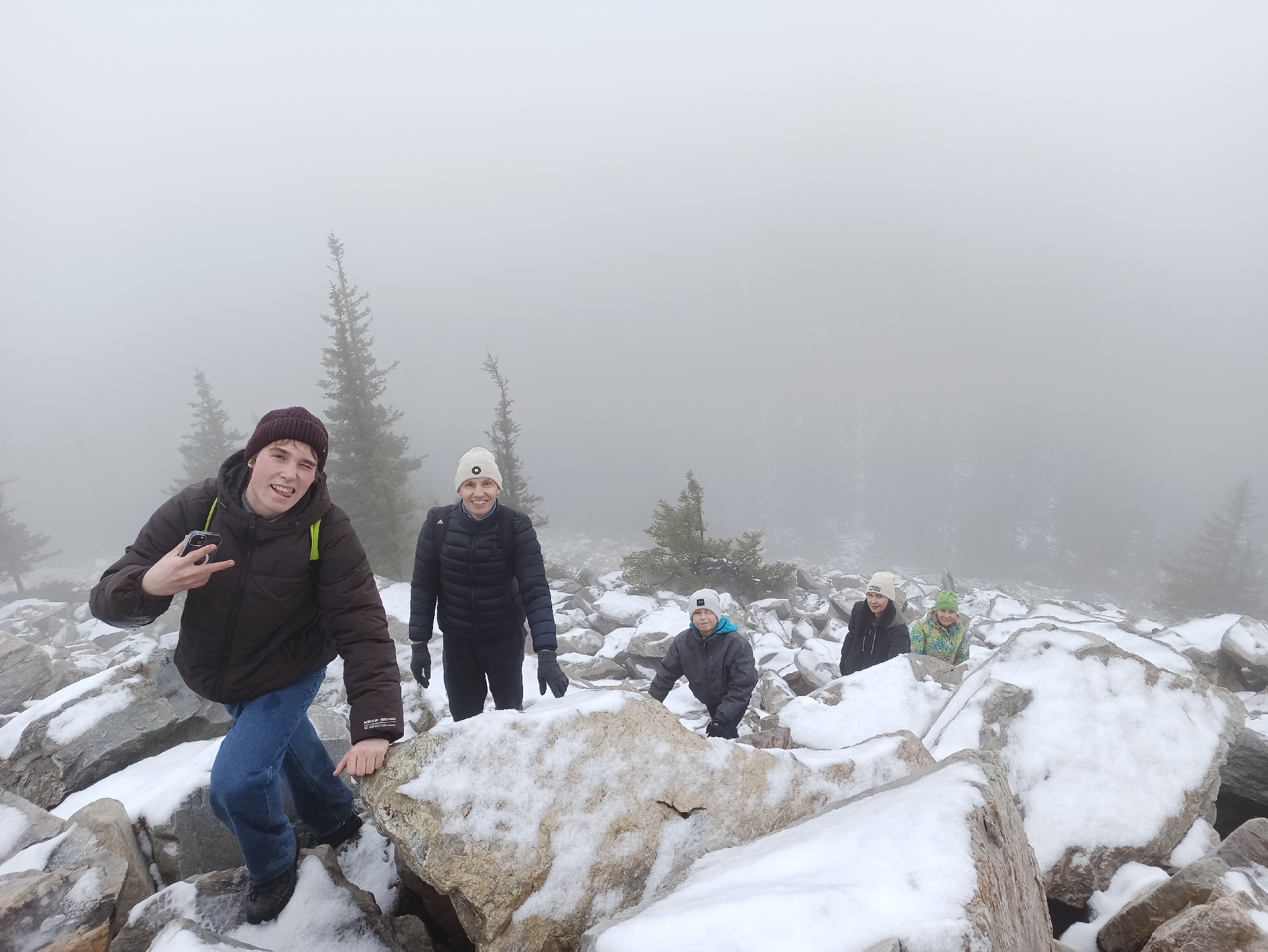 A group of five people are hiking in a rocky, snowy environment, obscured by heavy fog. The person in the foreground on the left is a young man wearing a dark maroon beanie, a brown puffer jacket with yellow accents, and blue jeans. He is stepping down from a rock, extending a peace sign with his left hand and holding a phone in the same hand. Behind him to the right is a person wearing a white beanie and a dark-colored puffer jacket, looking at the camera and smiling. Behind them is another person wearing a gray jacket and a white hat, crouching down on the rocks. Further back and to the right are two more people, also wearing winter hats and jackets. The ground is covered in large rocks, partially covered in snow. Pine trees are visible in the background, shrouded in fog. The overall scene suggests a mountain hike in cold, foggy weather. The location of Ziuratkul', Russia, is not directly discernible from the visual information provided.