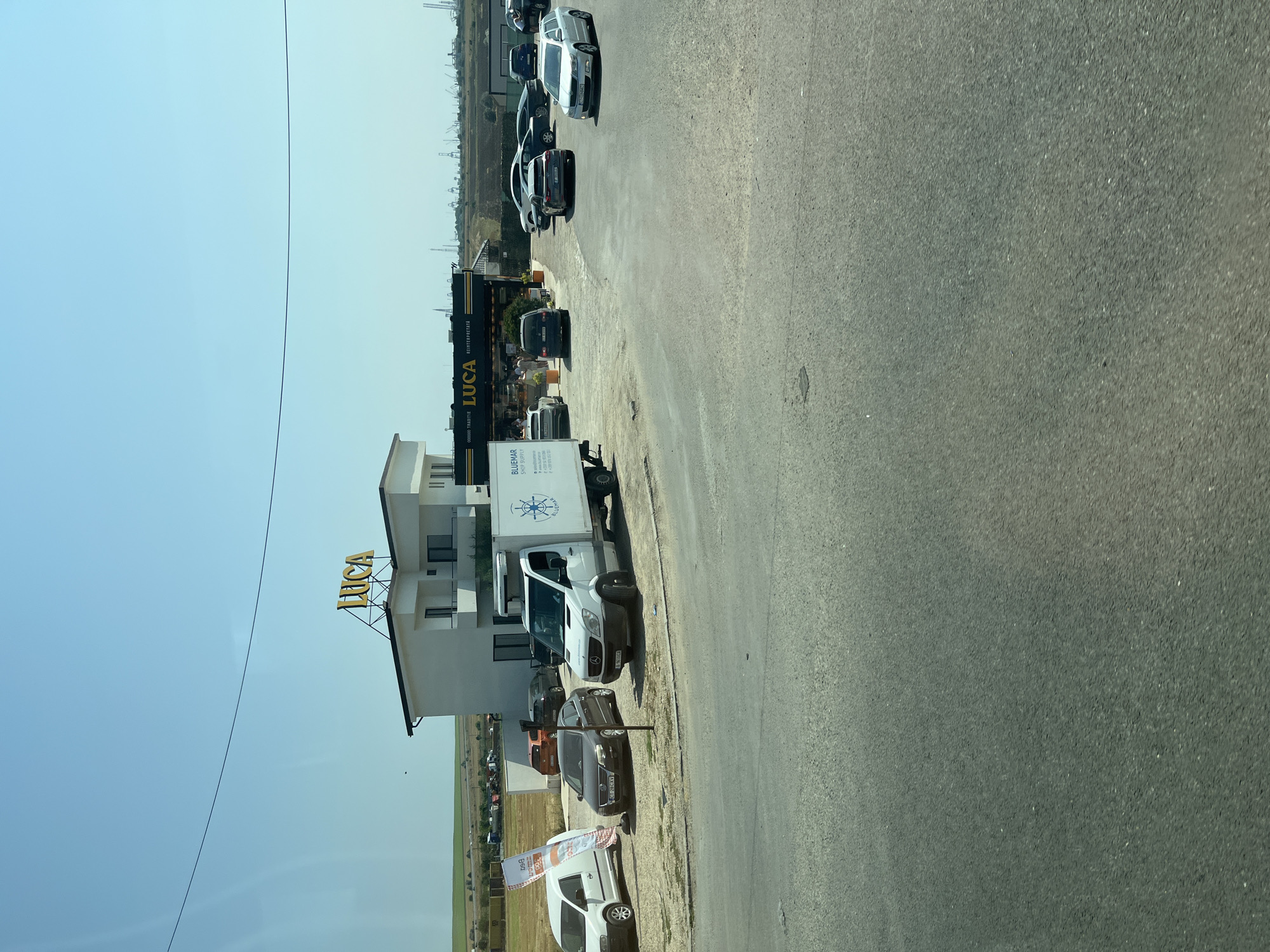 In Lazu, Romania, on a sunny day, a modern white building with a yellow "LUCA" sign is situated adjacent to a road. In front of the building, several cars are parked. A white delivery truck with "BLUEMAR" branding is parked on the right side of the road. A white van is parked to the left of the building, near a white banner. The background shows a flat landscape with fields and a distant industrial area with cranes. The road in the foreground is paved and appears to be a two-lane street. There are no visible people interacting in the scene. The overall impression is a commercial area on the outskirts of a town.