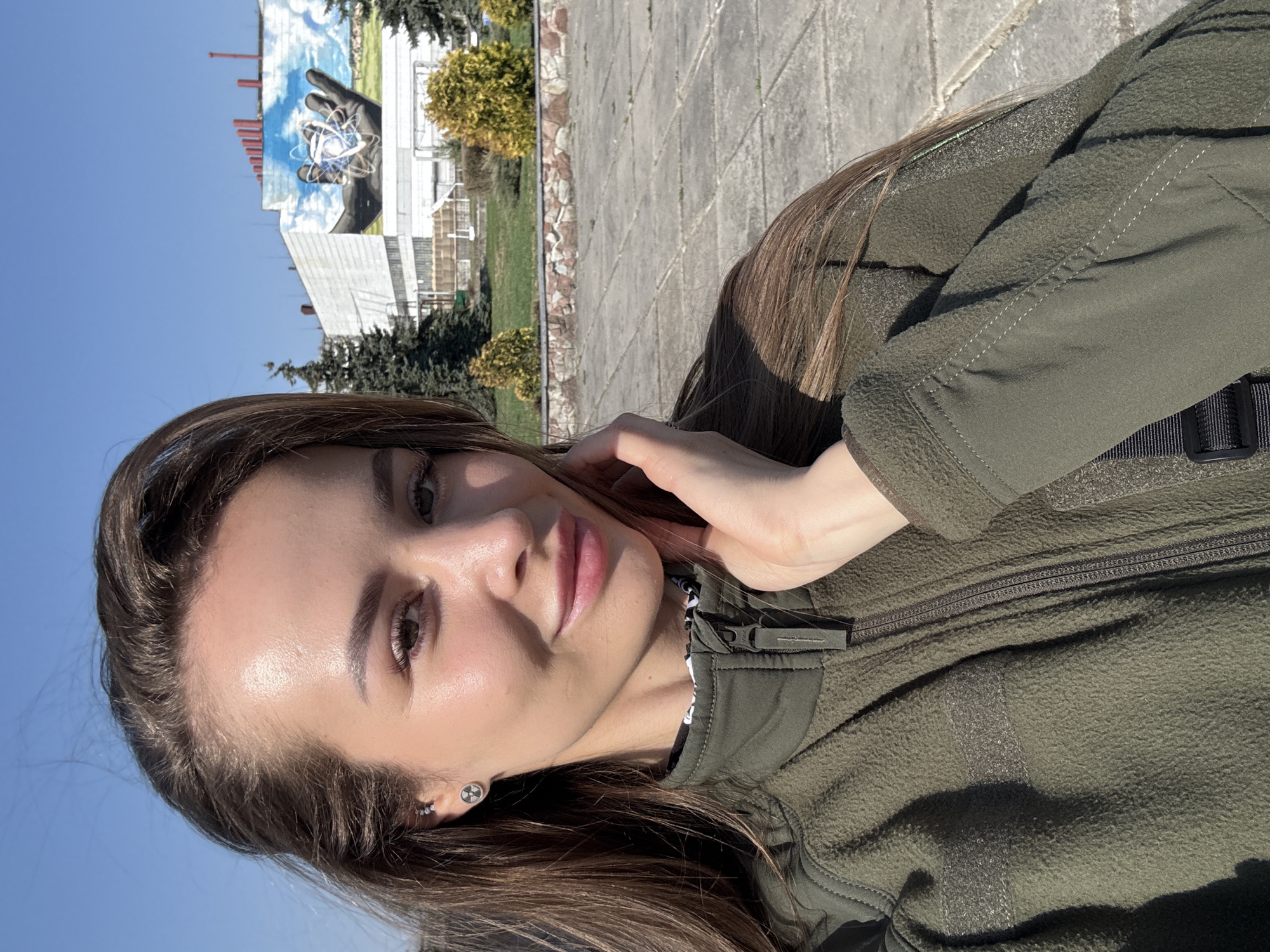 A woman is standing outside in what appears to be the city of Pryp'yat', Ukraine. She is wearing a dark green jacket and has dark brown hair. Her hand is touching her face, and she is looking directly at the camera. In the background is a building with a mural depicting a hand holding an atom. The weather is sunny, with a clear blue sky. The ground is paved with large stone tiles. Some trees and shrubs are also visible.