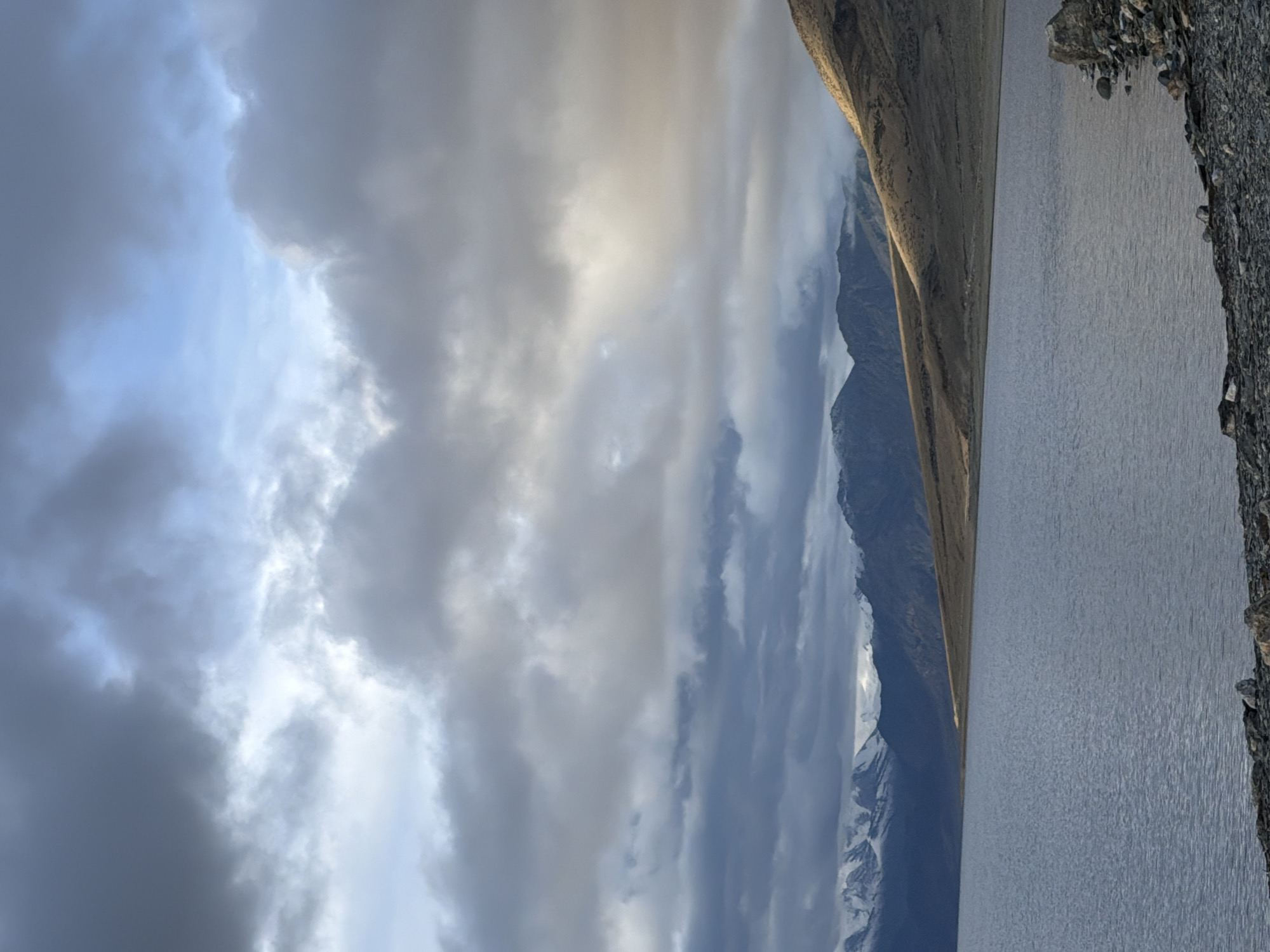This is a landscape photograph of a large, still body of water in front of a mountain range under a cloudy sky. The mountains in the background are dark and rugged, with some snow-capped peaks visible on the left side. The right side of the mountain range is illuminated by sunlight breaking through the clouds, creating a dramatic contrast. The water in the foreground is a muted blue-gray, with small ripples on its surface. The shore in the foreground is covered with dark, gray rocks and pebbles. The sky is filled with heavy, gray clouds, but there are patches of blue sky and shafts of sunlight visible, suggesting the weather is dynamic. The overall mood of the image is serene and majestic, with a sense of vastness and natural beauty. The location is identified as an unknown city in India.