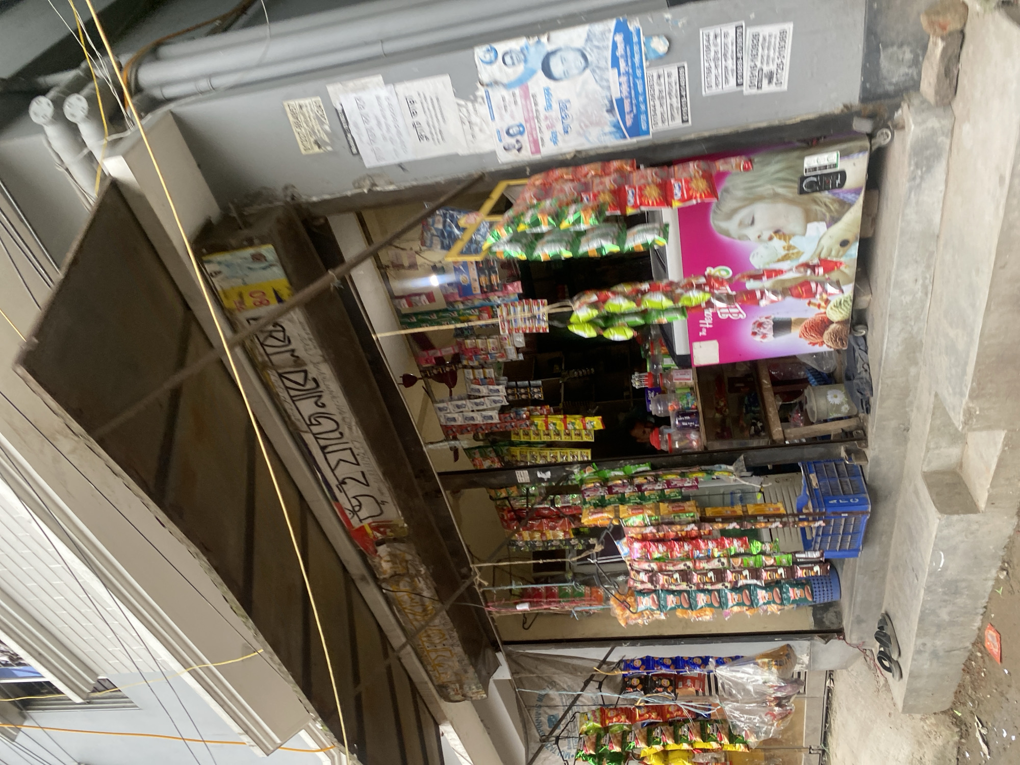 The image depicts a small street-side shop in Dhaka, Bangladesh. The shop is stocked with various snacks and food items, displayed on shelves and hanging from the ceiling. A large pink cooler with an advertisement of a girl eating ice cream is prominently placed in front of the shop. Various posters with text in Bengali are affixed to the wall next to the shop entrance. A pair of flip-flops is placed on the concrete steps leading up to the shop. The overall scene suggests a typical urban marketplace setting in Bangladesh.