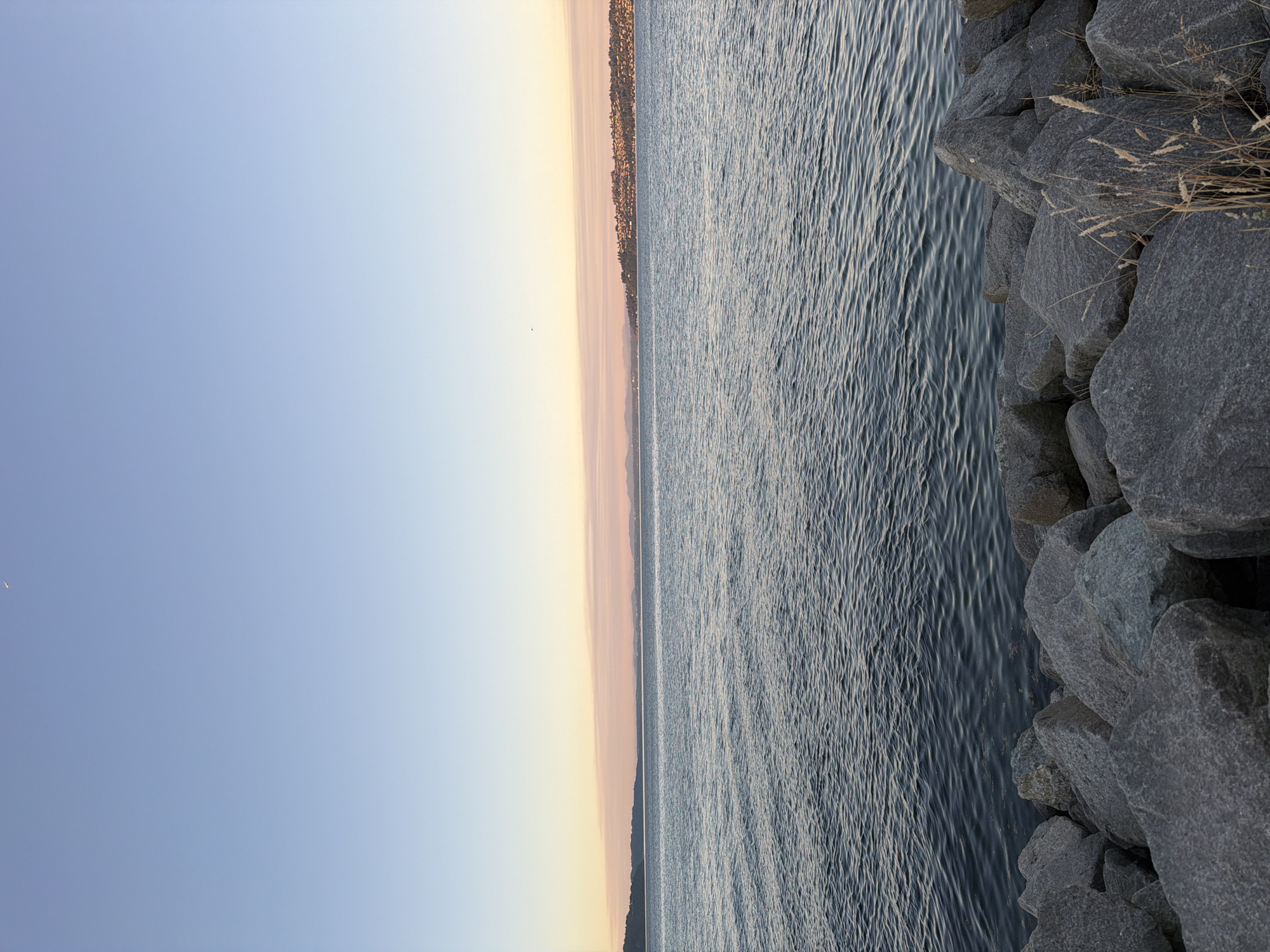 A wide-angle, eye-level shot captures a serene body of water against a soft, gradient sky, likely during dusk. The water, a deep blue-grey, is textured with gentle ripples, indicating a calm surface. In the foreground, a dense arrangement of large, grey rocks forms a natural barrier. These rocks vary in size and shape, with some appearing rough and unpolished. Amongst the rocks, dry, wispy grass adds a touch of muted color.
The middle ground is dominated by the vast expanse of water, which meets a distant horizon. Along this horizon, a sliver of land is visible, featuring a dense cluster of buildings that suggest a city or town nestled against a backdrop of rolling hills or mountains, partially obscured by a faint haze. The sky transitions from a pale blue at the top of the frame to soft hues of pink and orange near the horizon, a characteristic of sunset or sunrise. A tiny, almost imperceptible crescent moon is visible in the upper left corner of the sky. A few distant, faint streaks of clouds run horizontally across the sky, adding subtle texture. The overall impression is one of natural tranquility with a hint of distant civilization. The scene is devoid of any human presence or overt activity.