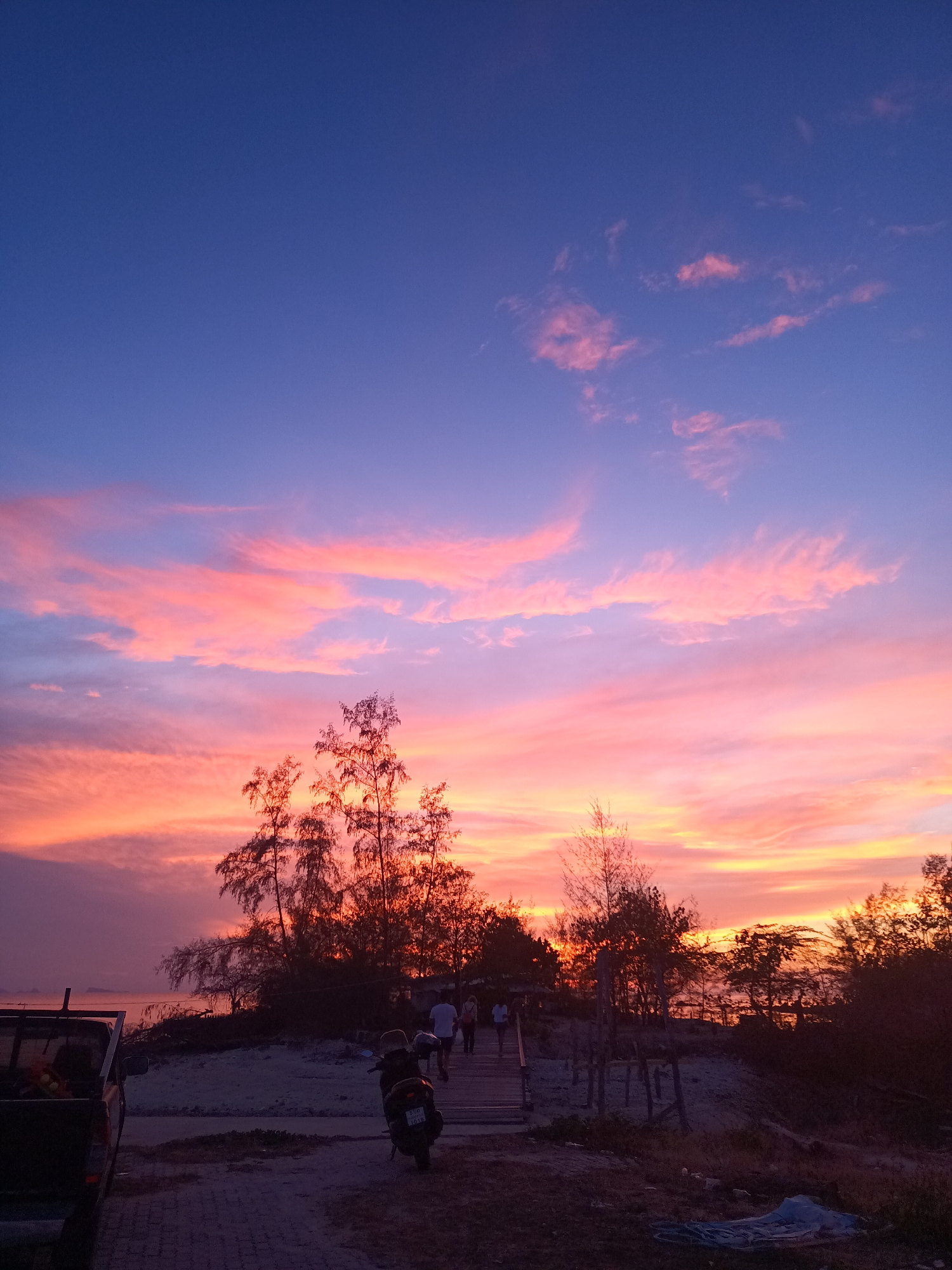 The image shows a sunset scene in Baan Sri Thanu, Thailand. The sky displays a gradient of colors, with deep blue at the top transitioning to pink and orange hues near the horizon. Silhouetted trees are in the foreground, along with a group of people walking on a wooden path. A parked motorcycle and a truck are visible on the left side of the image. The ground is a mix of paved surface and sand.