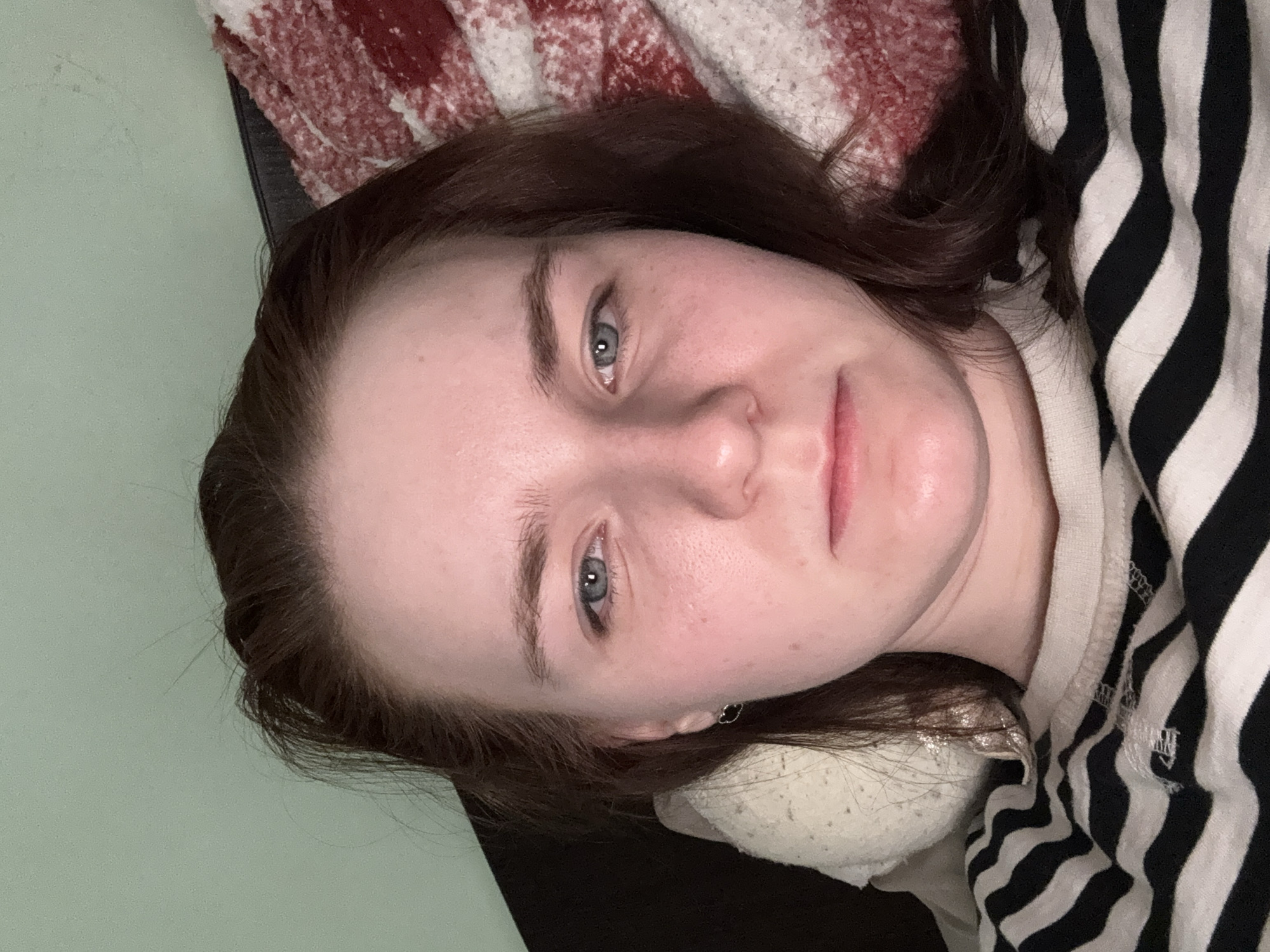A close-up, low-angle shot depicts a woman lying down. Her head rests on a black surface, possibly a couch or bed, covered with a red and white patterned blanket. She is wearing a black and white striped shirt. Her hair is brown, and her eyes are light blue. An earring is visible in her left ear. The background includes a pale green wall. The location is Unknown, Russia.