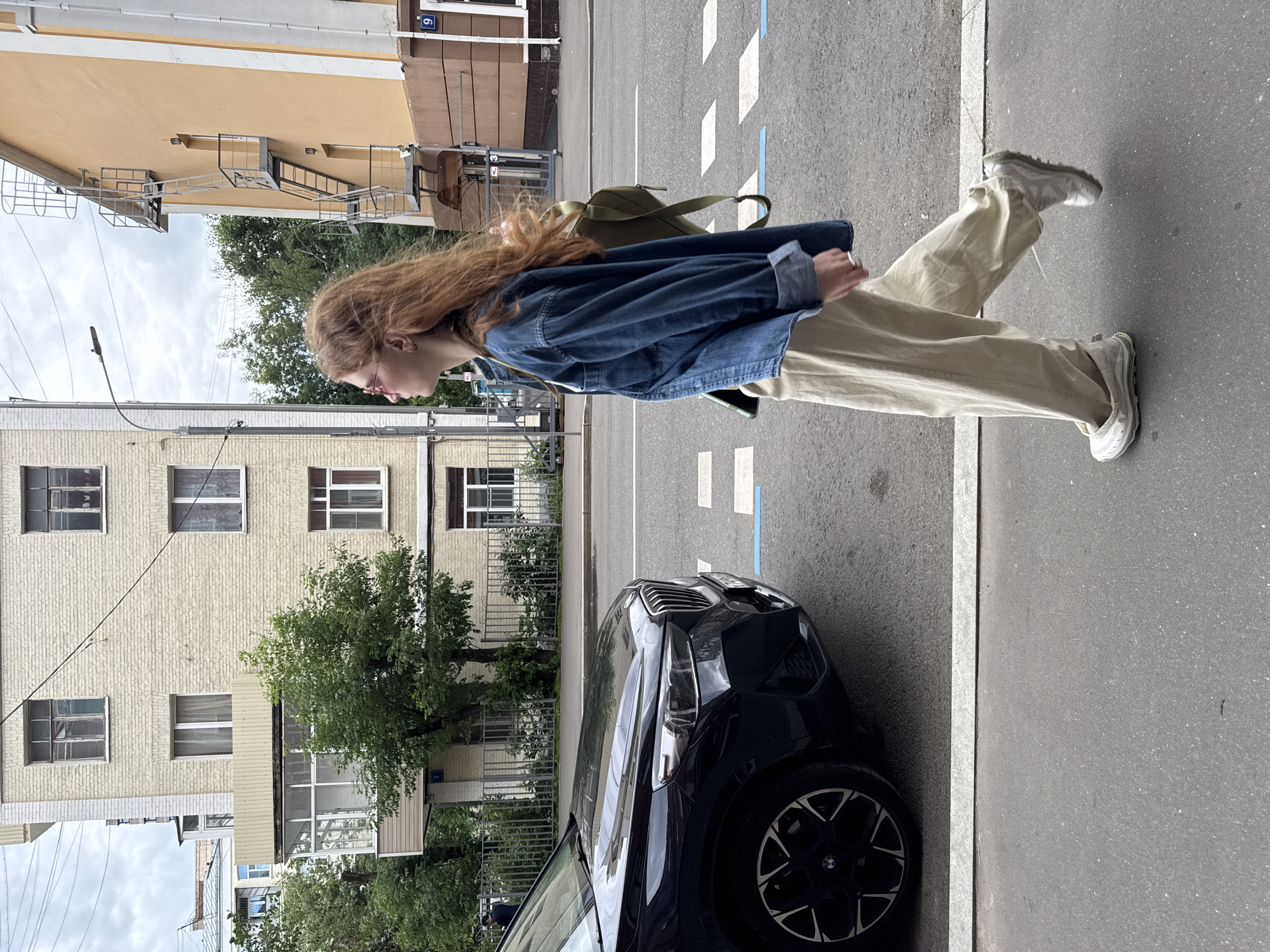 A young woman with long, reddish-blonde hair walks along a city street in Moscow, Russia. She is wearing a blue denim jacket, beige pants, and white sneakers, and has a green backpack slung over her shoulder. Her profile is visible as she looks down, and she appears to be holding a cigarette in her right hand. A black car is parked beside her on the street, which has pedestrian crossing markings. Buildings with yellow and beige facades, some with exposed brickwork and many windows, line the street. Trees with green foliage are visible between the buildings. The sky is overcast, suggesting a cloudy day. The scene captures a casual moment of urban life.