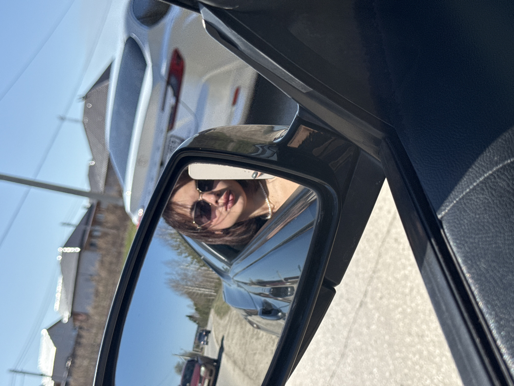 The image shows a side mirror of a car reflecting a woman with sunglasses in Лобаново, Russia. A white car is visible behind the side mirror. The setting seems to be a street with buildings and other cars present.