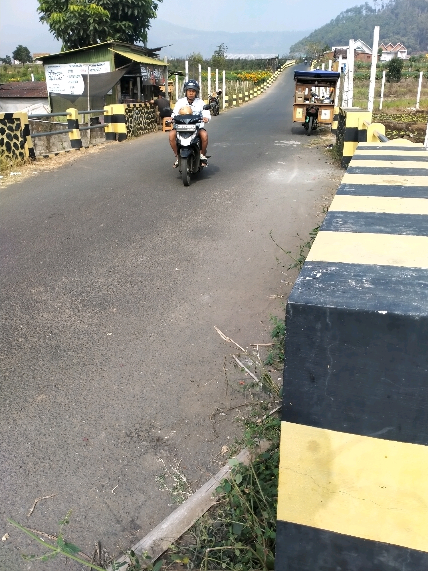 A man wearing a white shirt and a black helmet rides a black motorcycle on a paved road with yellow and black striped barriers on the side. The road appears to be in a rural or semi-rural area, possibly in Batu, Indonesia, given the style of the barriers and the presence of small shops and greenery. In the background, there is a small building with a green roof and signage. Further in the distance, hills and houses are visible under a hazy sky. To the right of the road, there is a cart attached to a motorcycle, and more yellow and black barriers. The overall scene depicts a typical roadside view in a tropical or sub-tropical region.